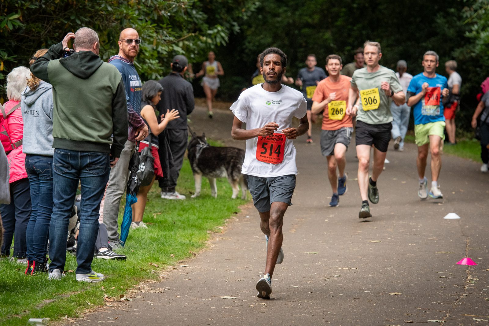 A man wearing a white shirt and red race bib 514 runs on a park path during a race, followed by other runners. Spectators and a dog stand on the grass beside the path, watching and cheering.
