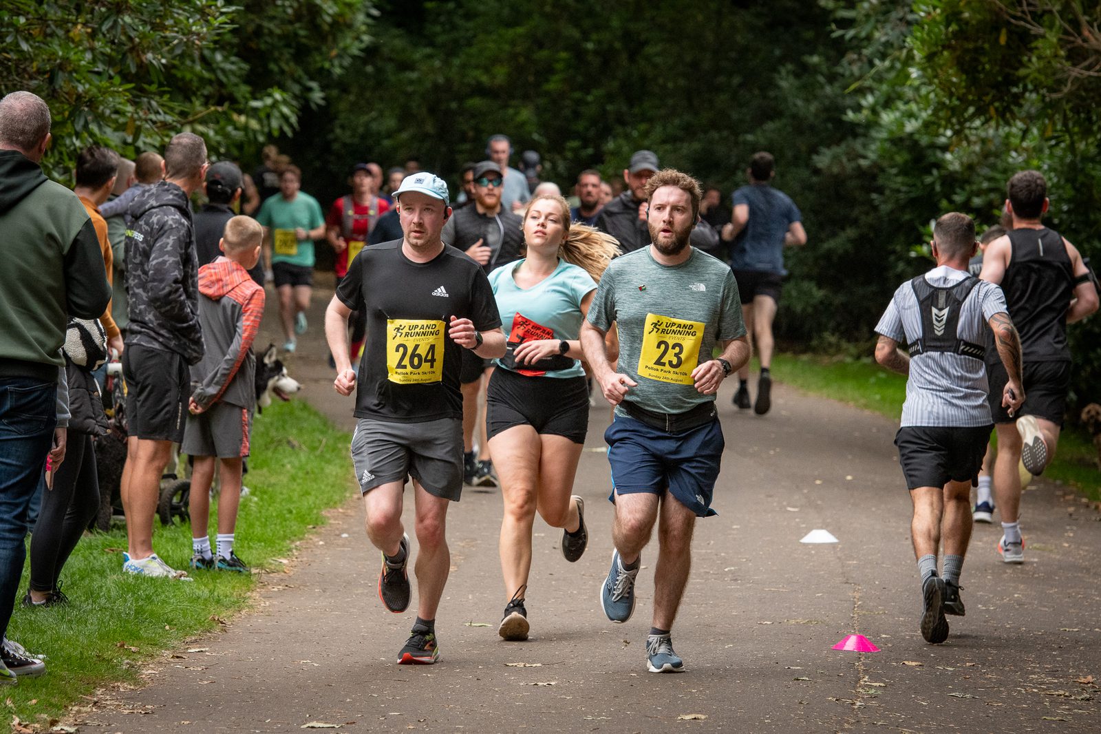 Three runners with race bibs 264, 4, and 23 jog closely together on a park path during a race, surrounded by other runners and spectators in a green, wooded setting.