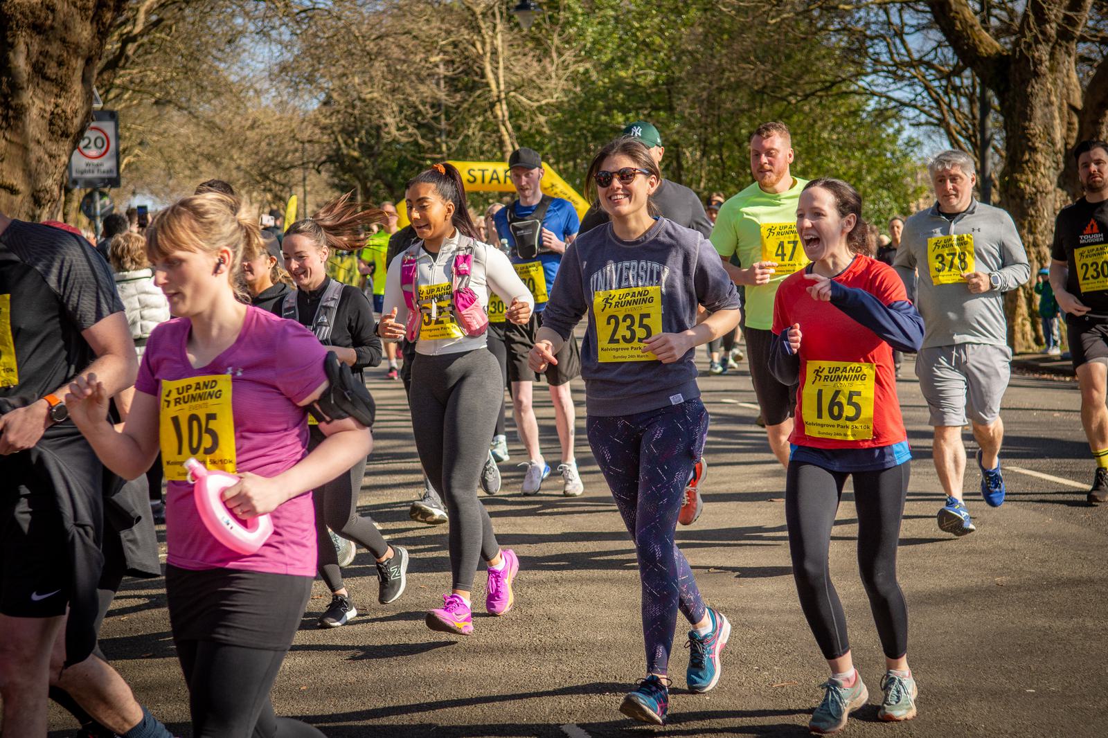 A group of runners participating in a race. Various people, dressed in athletic attire, jog down a tree-lined street, some smiling and chatting. Bib numbers, including 105, 165, 235, and 47, are visible. Bystanders watch from the background.
