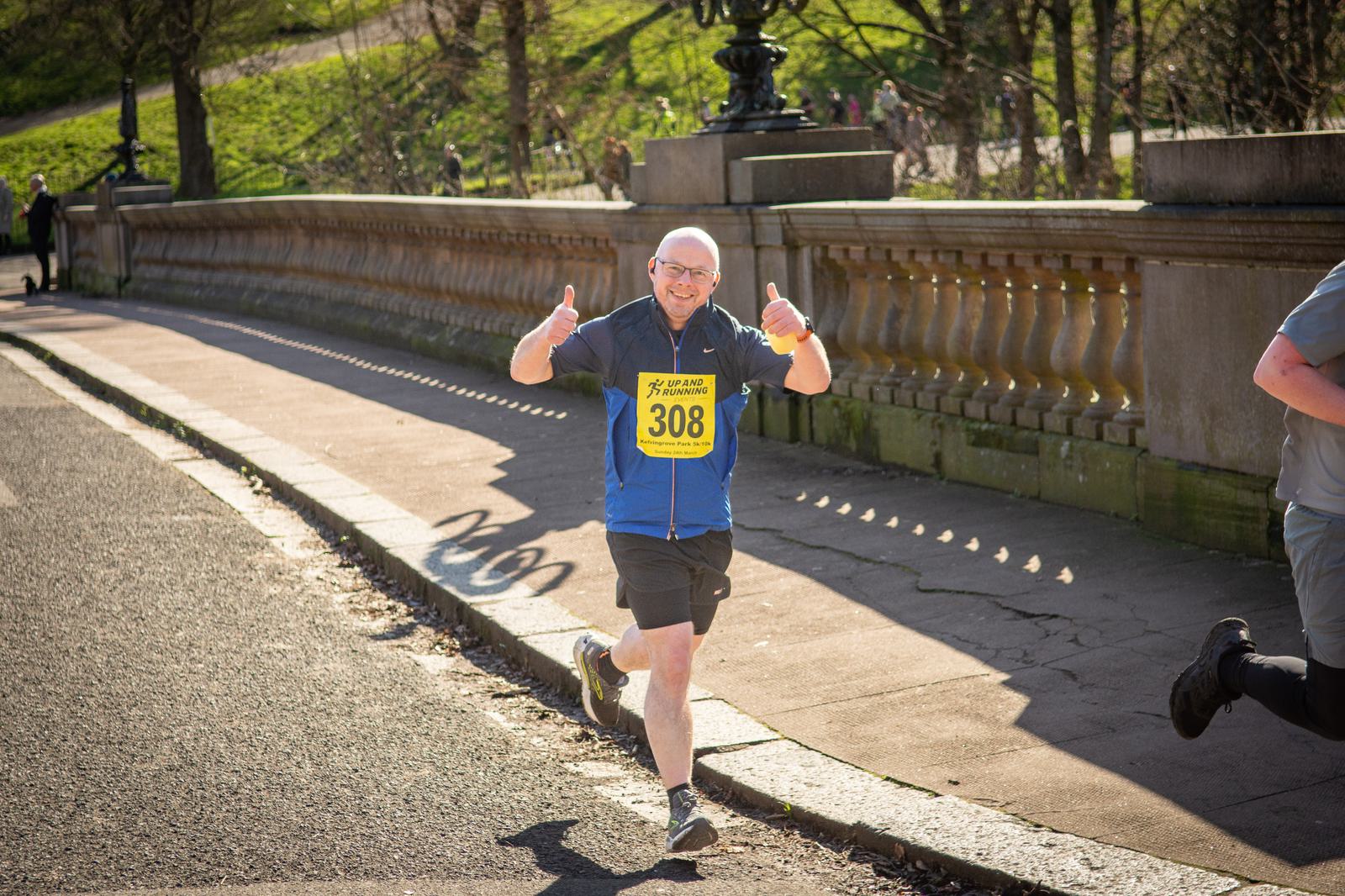 A man with a shaved head wearing glasses, a blue and black running outfit, and a race bib number 308 is smiling and giving a thumbs-up as he runs outdoors on a paved path beside a stone railing, with greenery and people in the background.