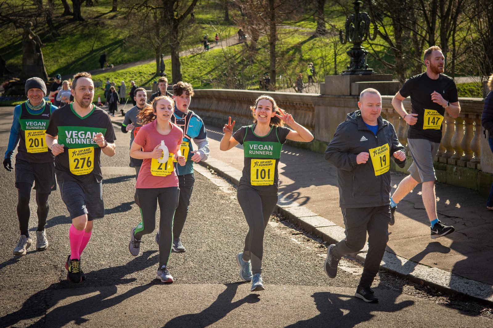 A group of people jogging in a marathon on a sunny day. Some wear numbered bibs, and two participants in the foreground wear green "Vegan Runners" shirts. They look cheerful; a woman in the center is making a peace sign. Others behind them and to the sides also jog.