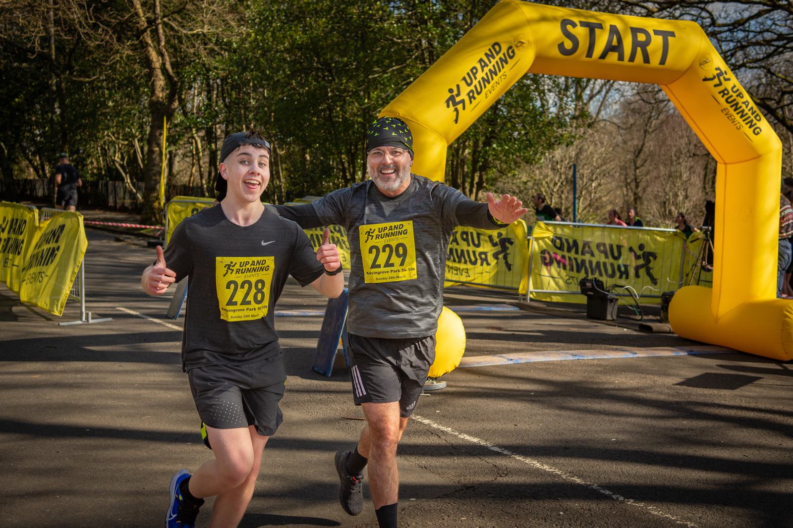 Two runners wearing race bibs 228 and 229 smile and gesture enthusiastically as they pass under a yellow starting archway during a race. Both are dressed in athletic gear, with trees and race barriers in the background.