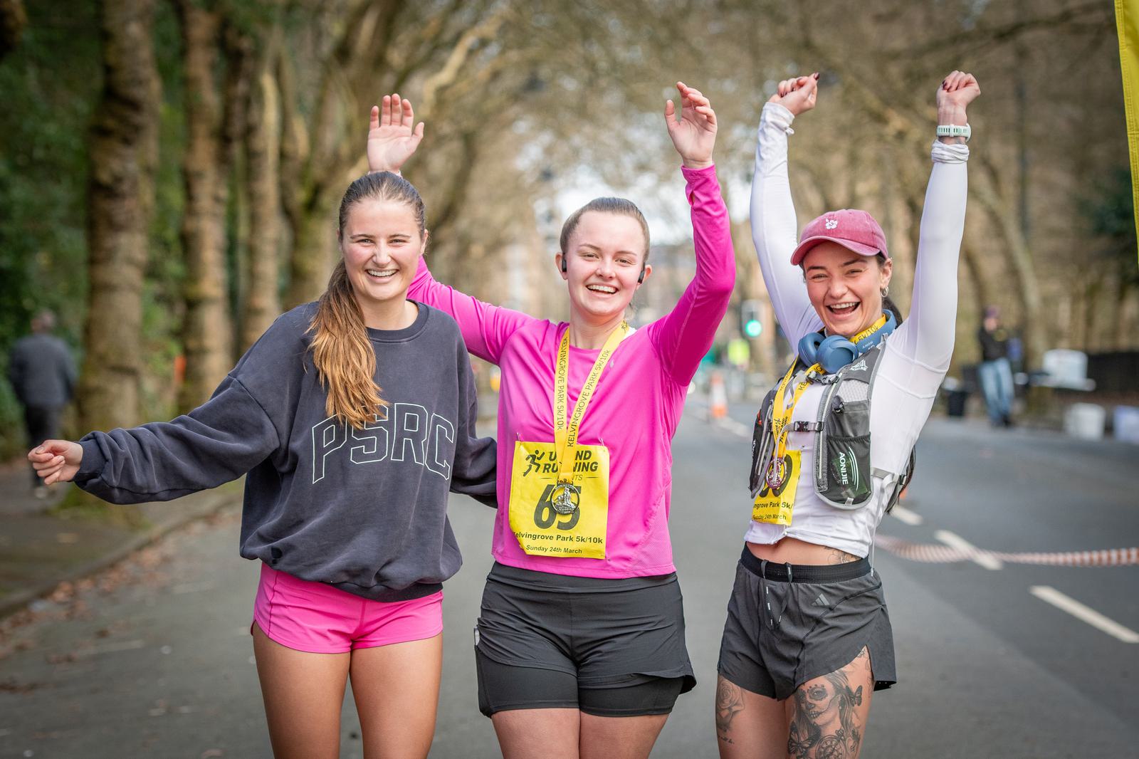 Three women stand together on a road, smiling and raising their arms in celebration. They are dressed in athletic wear and have race bibs pinned to their shirts. Trees line the background, indicating an outdoor setting, likely post-race or during a fun run.
