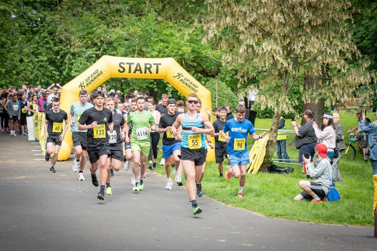 A group of runners starts a race at a park, passing under a yellow "START" arch, with spectators and photographers on the side. The runners wear numbered bibs and athletic clothing. Green trees surround the path.