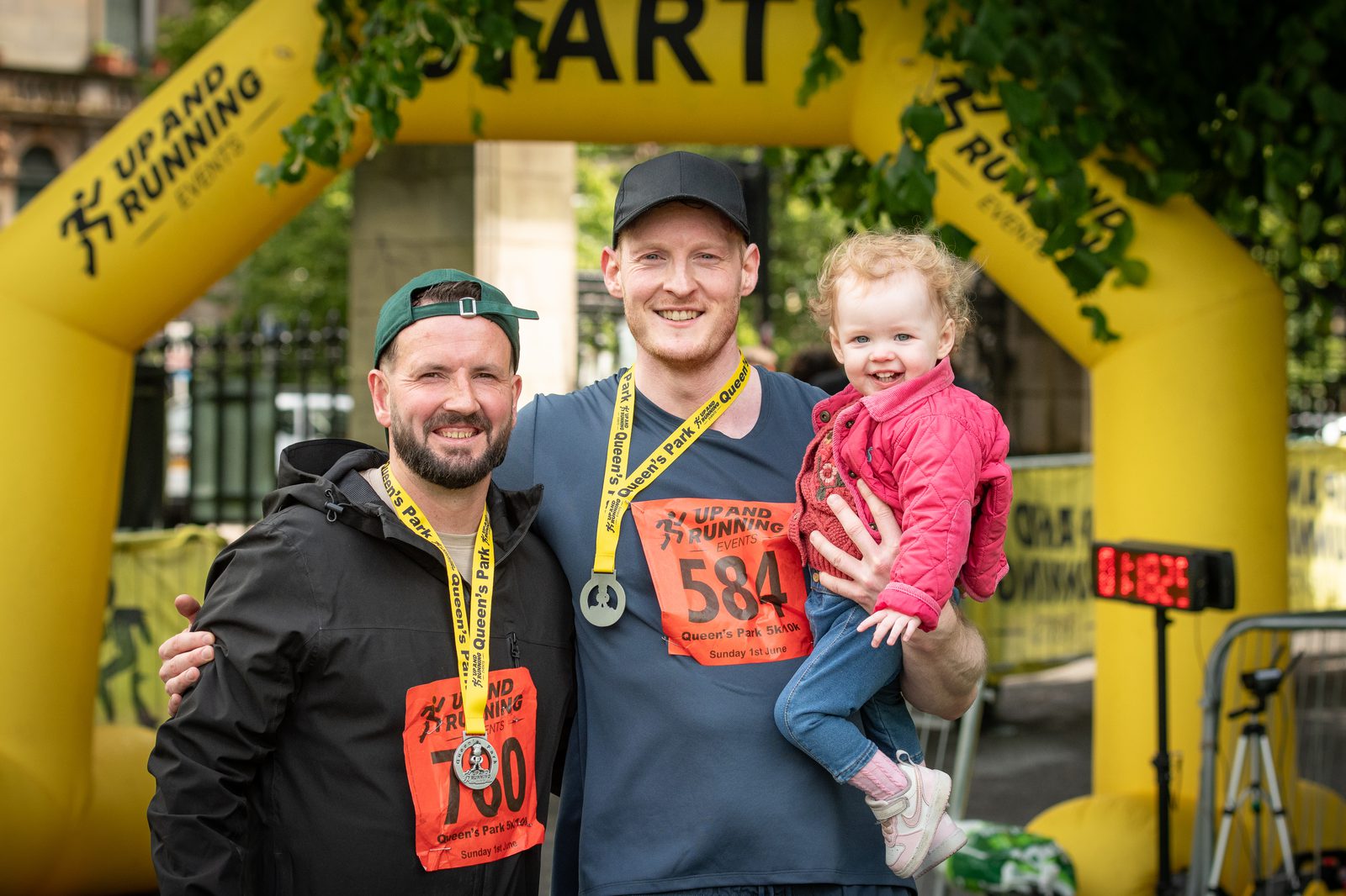 Two men with race bibs and medals smile at the camera under a yellow "Up & Running" race arch. One man holds a toddler in a pink jacket. Trees and race equipment are visible in the background.
