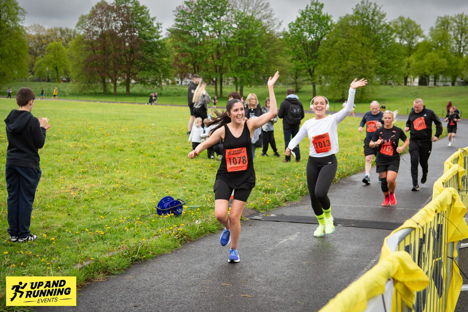 Two women, wearing race bibs 1078 and 1013, cheerfully cross the finish line of a race in a park. They are both smiling and raising their arms in celebration. Behind them, other participants are also finishing. A yellow race banner and "Up and Running Events" logo are visible.