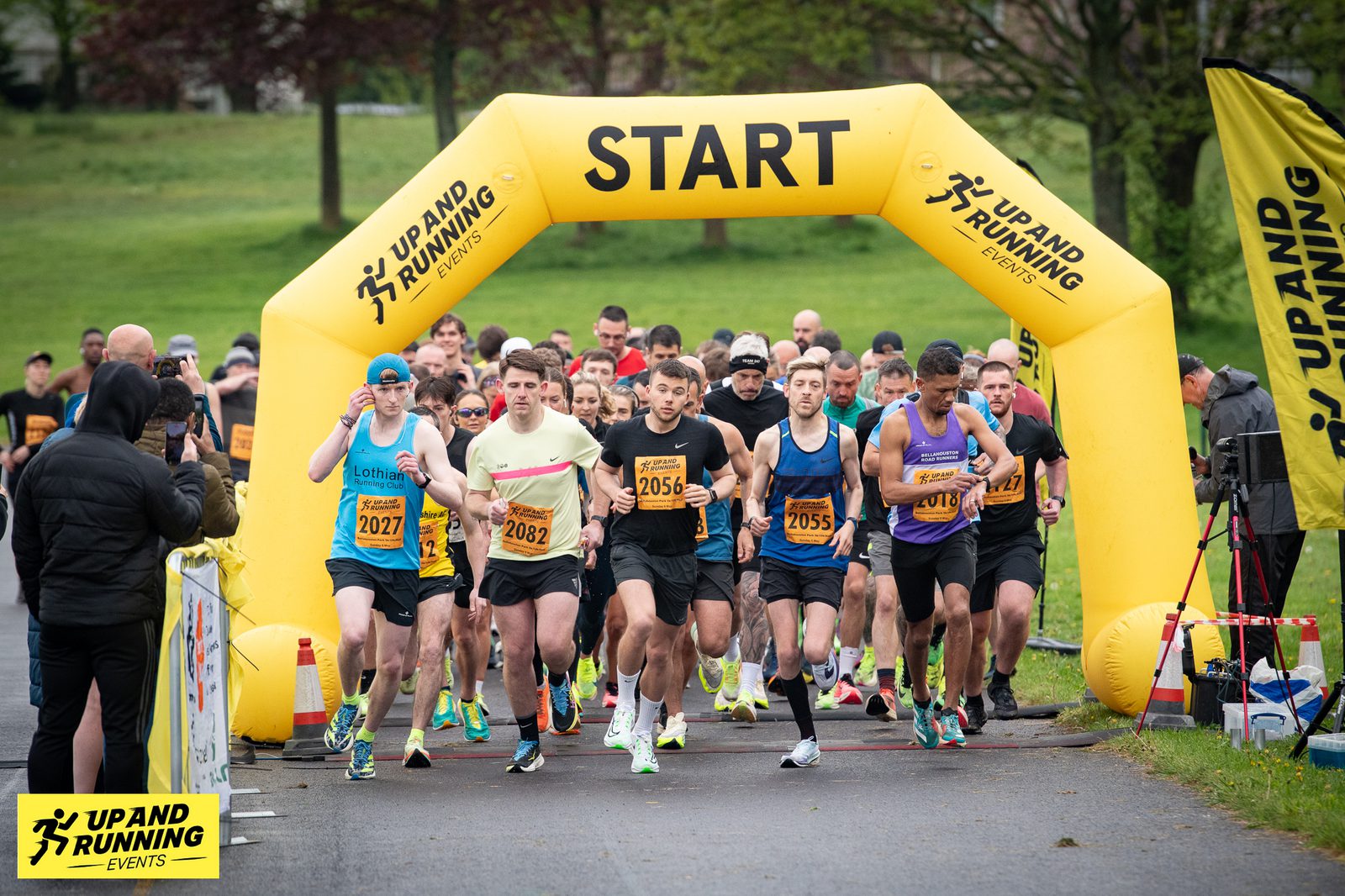 A group of runners, wearing various athletic clothing and race bibs, begins a race by running through a yellow inflatable start arch with "START" written on it. The event is organized by "Up and Running Events," as indicated by the signage and banners. Trees and grass are visible in the background.