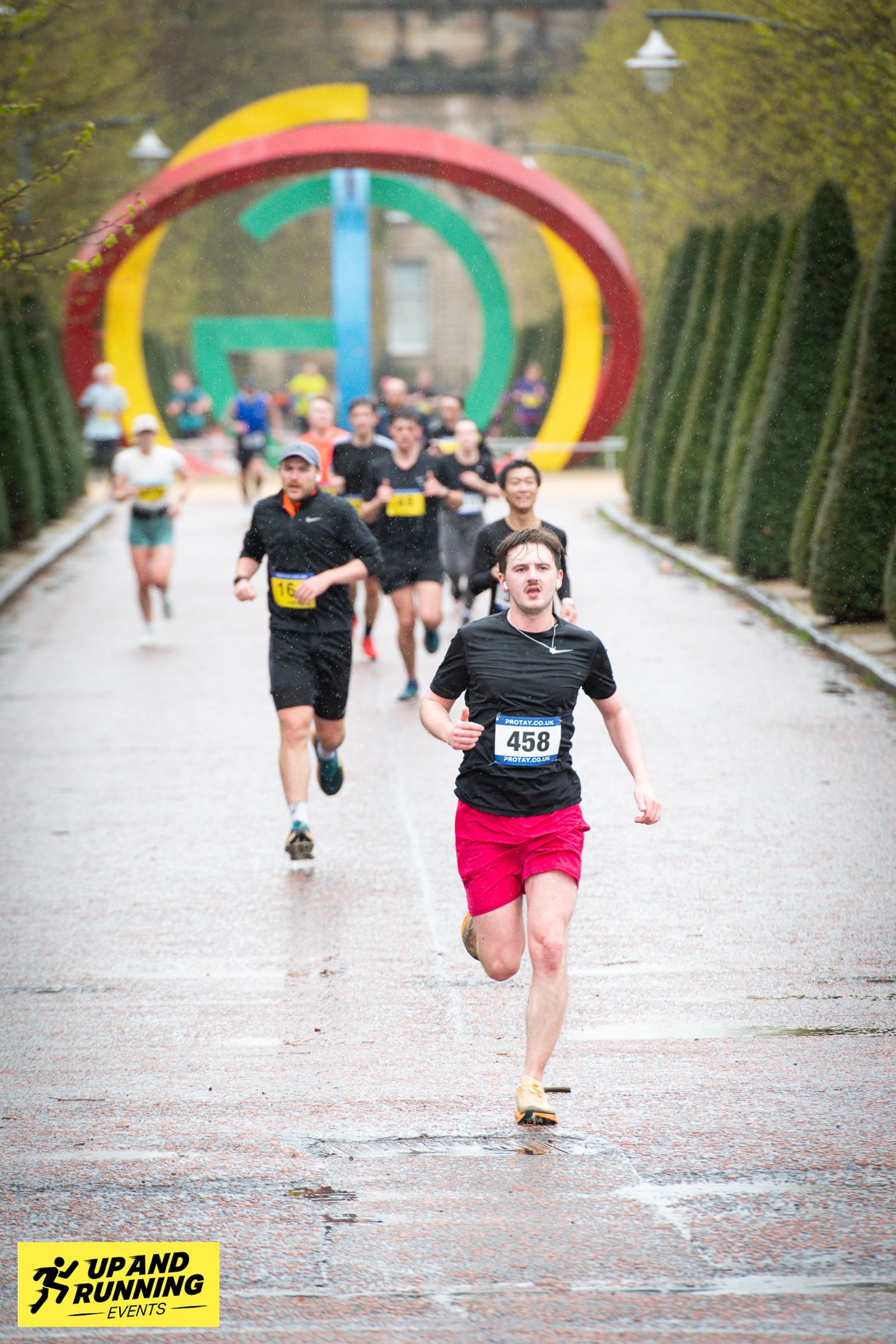 Runners in a race, with one person in red shorts leading on a rain-soaked street. A colorful archway is in the background. The road is lined with trees, and there's a logo for "Up and Running Events" in the foreground.