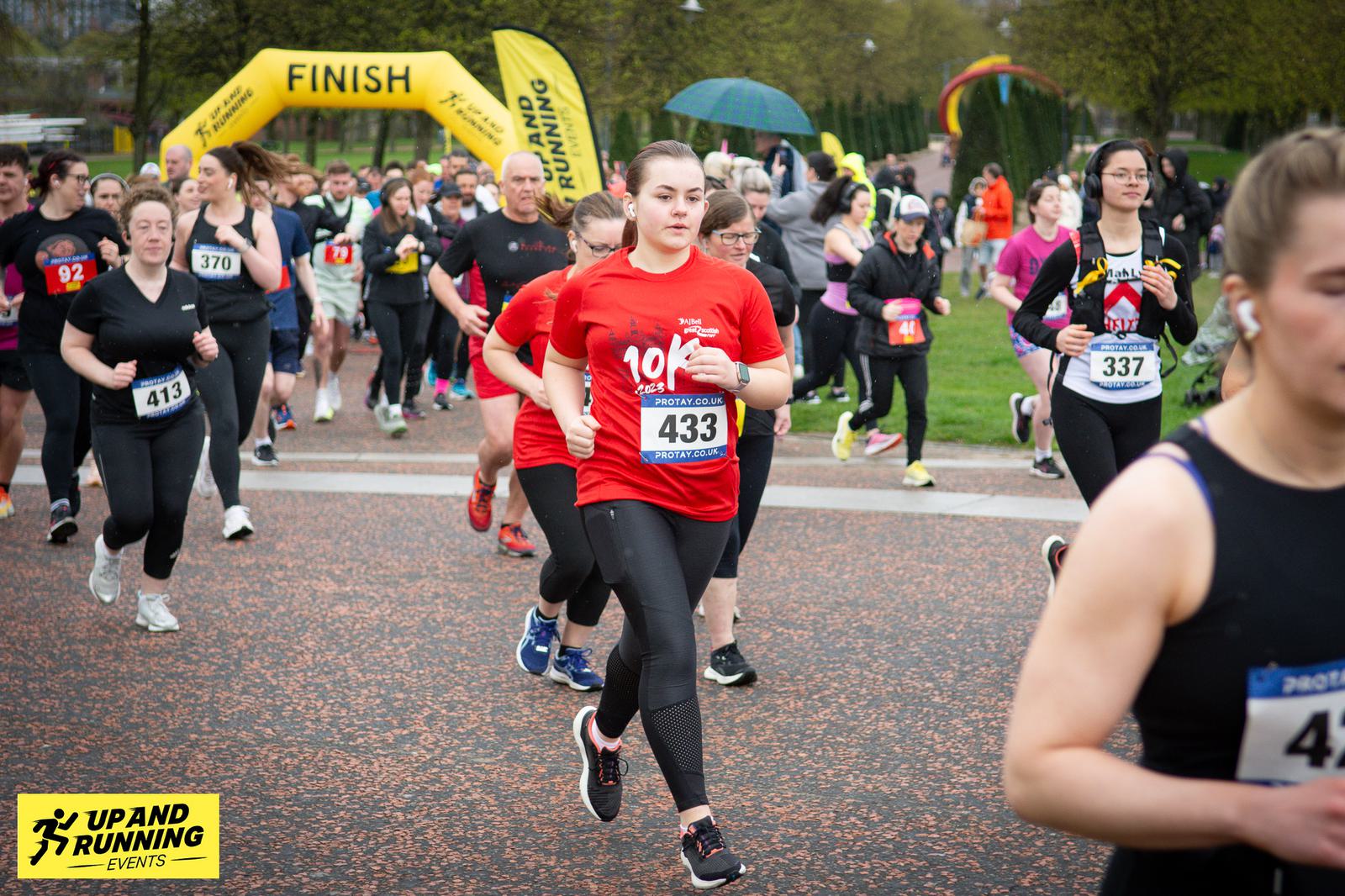 A group of runners participating in a race near the finish line. One runner in the foreground is wearing a red shirt with the number 433. The event is organized by "Up and Running Events," as shown by the banner.