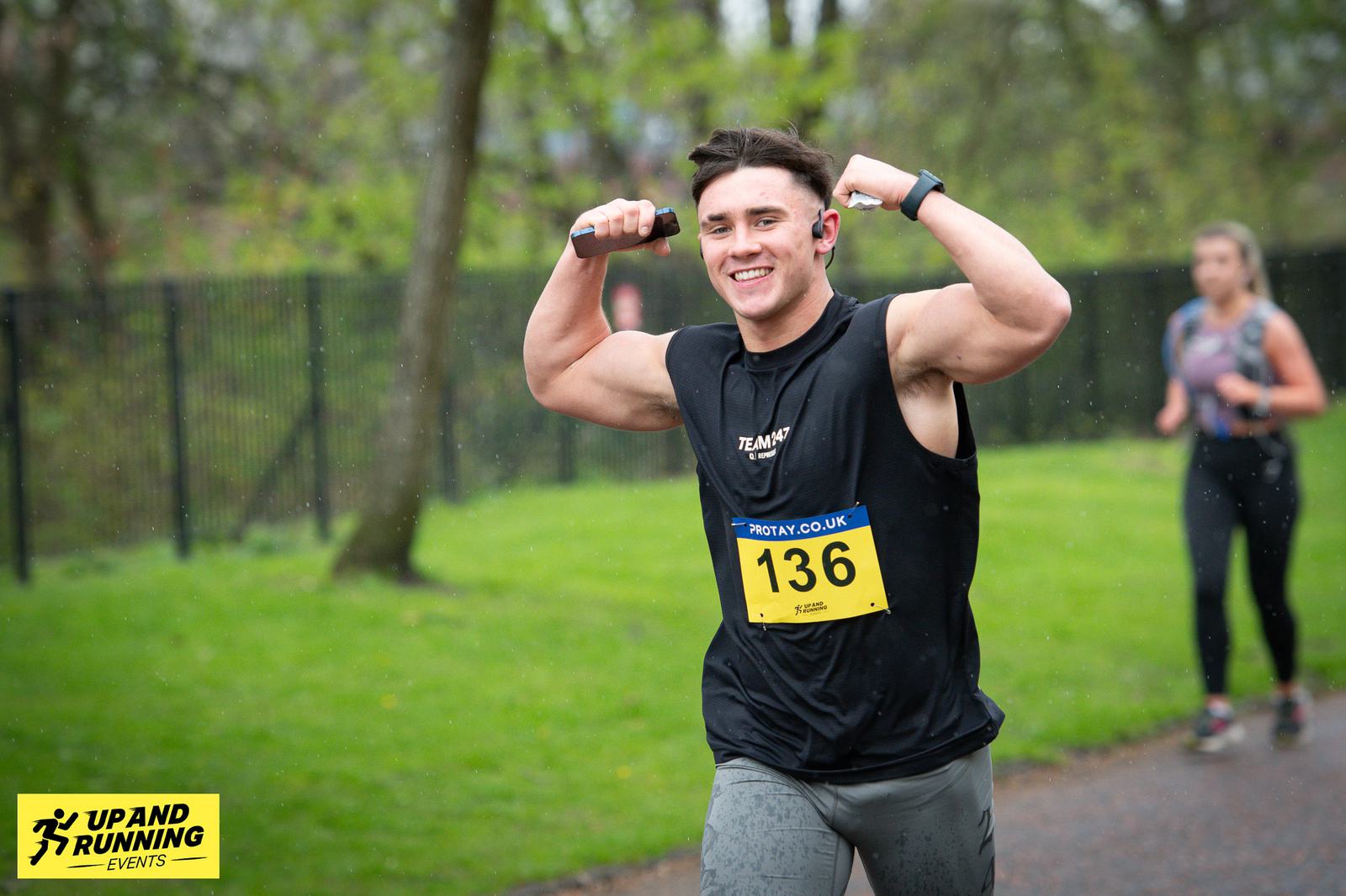 A smiling runner in a sleeveless black shirt flexes their arms while running a race in a park. They wear a yellow race number 136. A logo in the corner reads "Up and Running Events." Another runner is visible in the background.