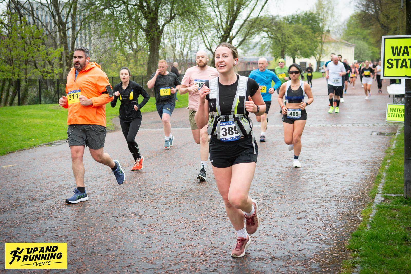 Runners participating in a race on a wet road surrounded by trees. The lead runner is smiling and wearing a black outfit with a bib number 338. Other runners follow, and a sign for a water station is visible in the background.