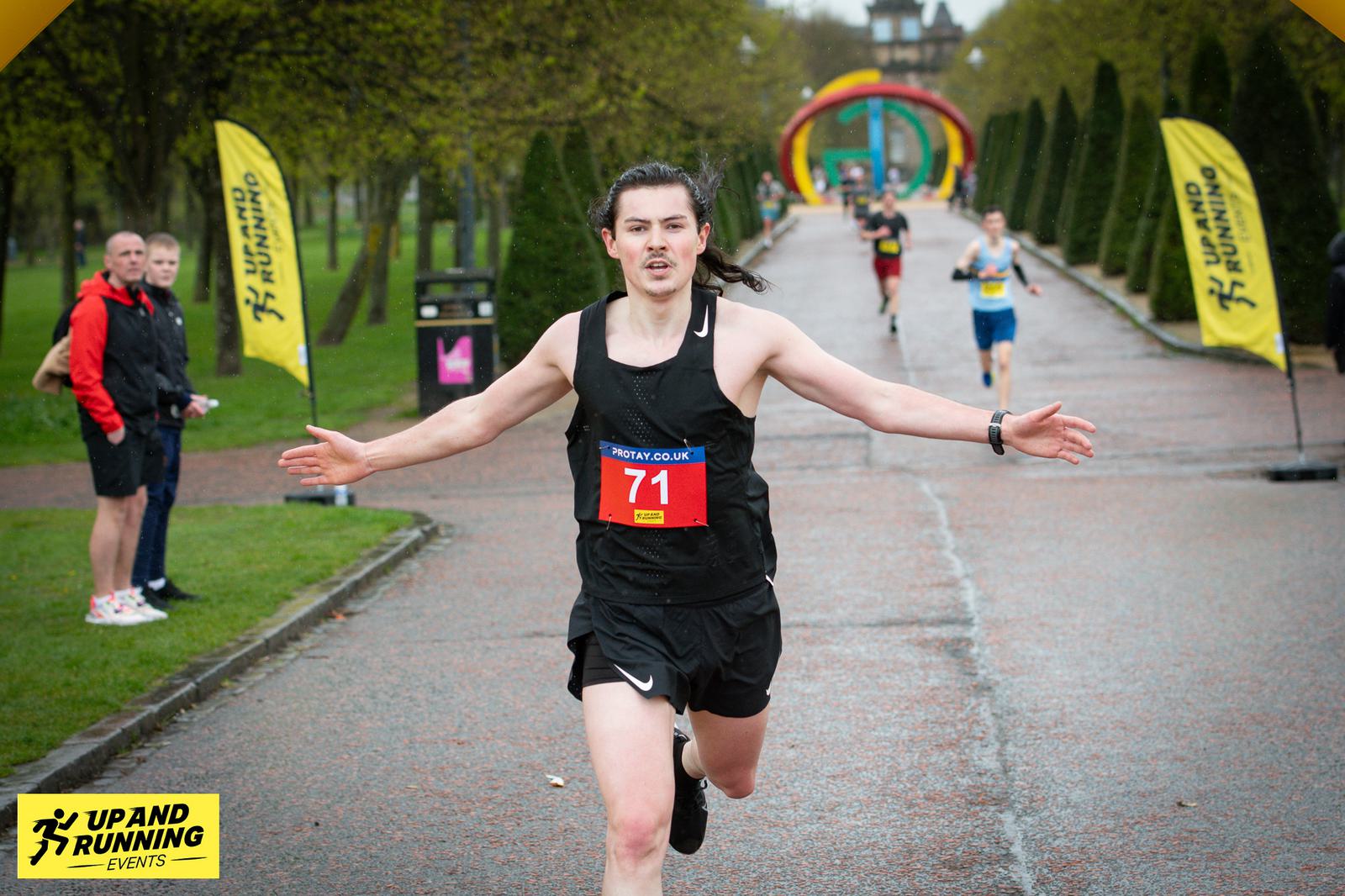 A runner wearing a black outfit and race bib number 71 crosses the finish line with arms outstretched. The background shows other runners, event flags, and a tree-lined path. The ground is wet, indicating recent rain. Signs read "Up and Running Events.