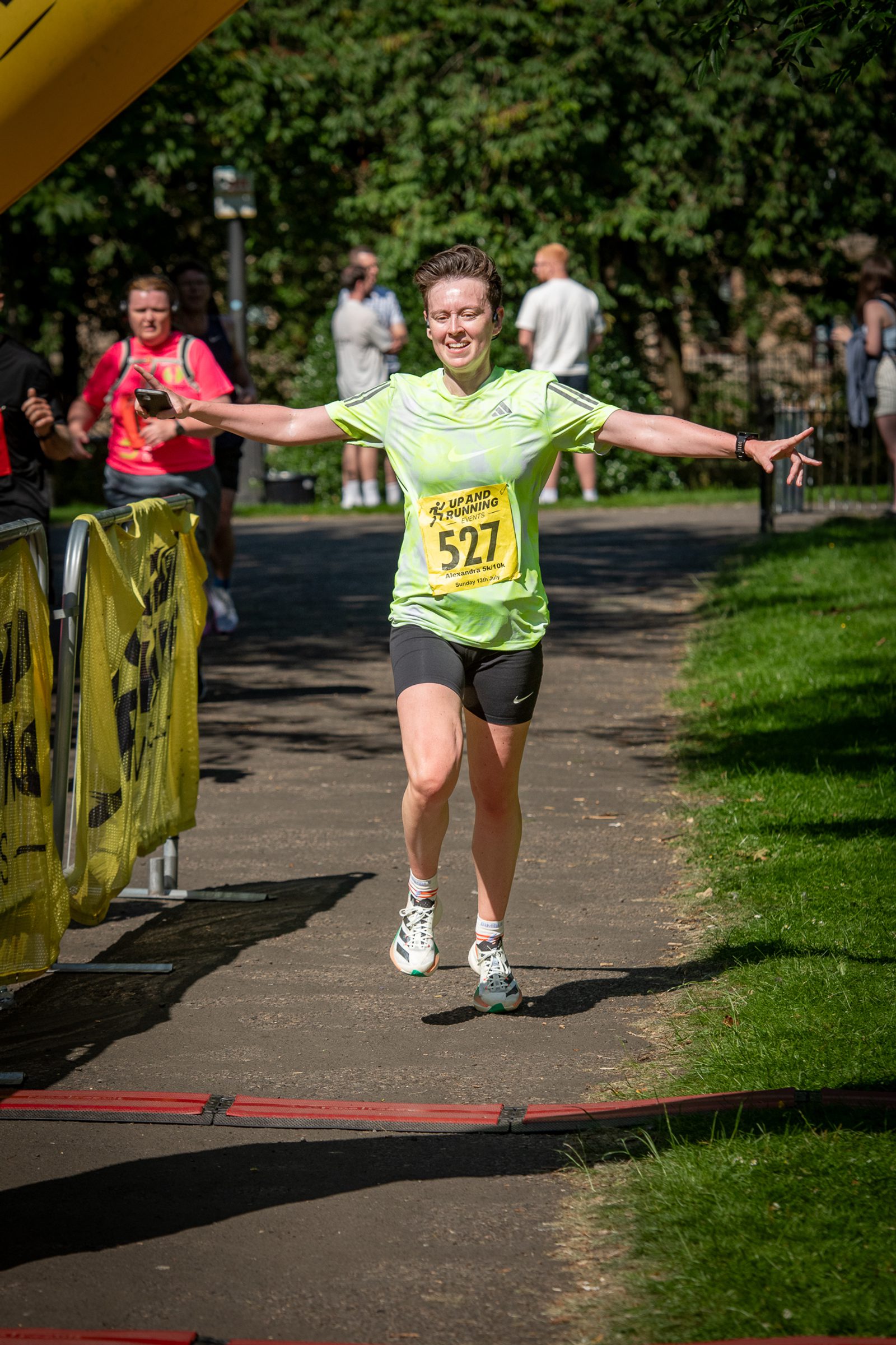 A runner wearing a green shirt, black shorts, and race bib 527 smiles and spreads their arms wide as they cross a finish line in a sunny park, with spectators and trees in the background.