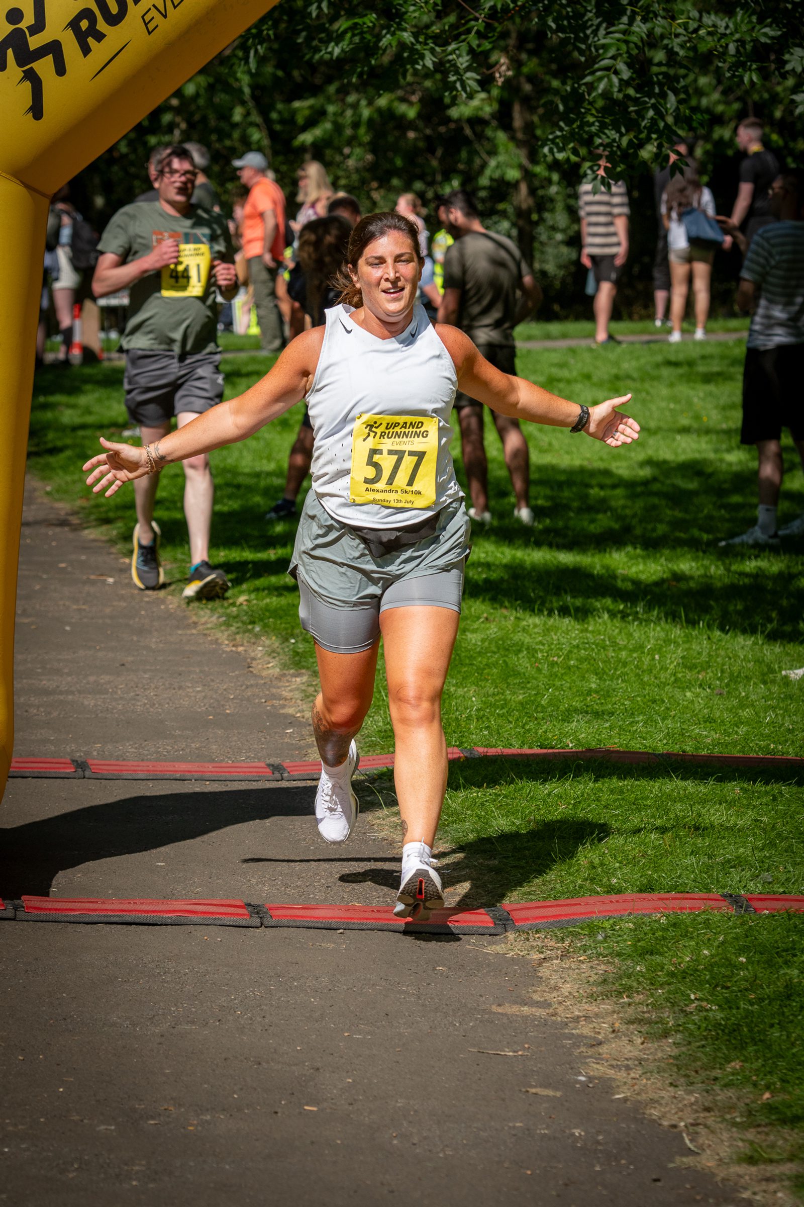 A woman wearing a race bib numbered 577 smiles with arms wide as she crosses a finish line in a park. Other runners and spectators are visible in the background on a sunny day.