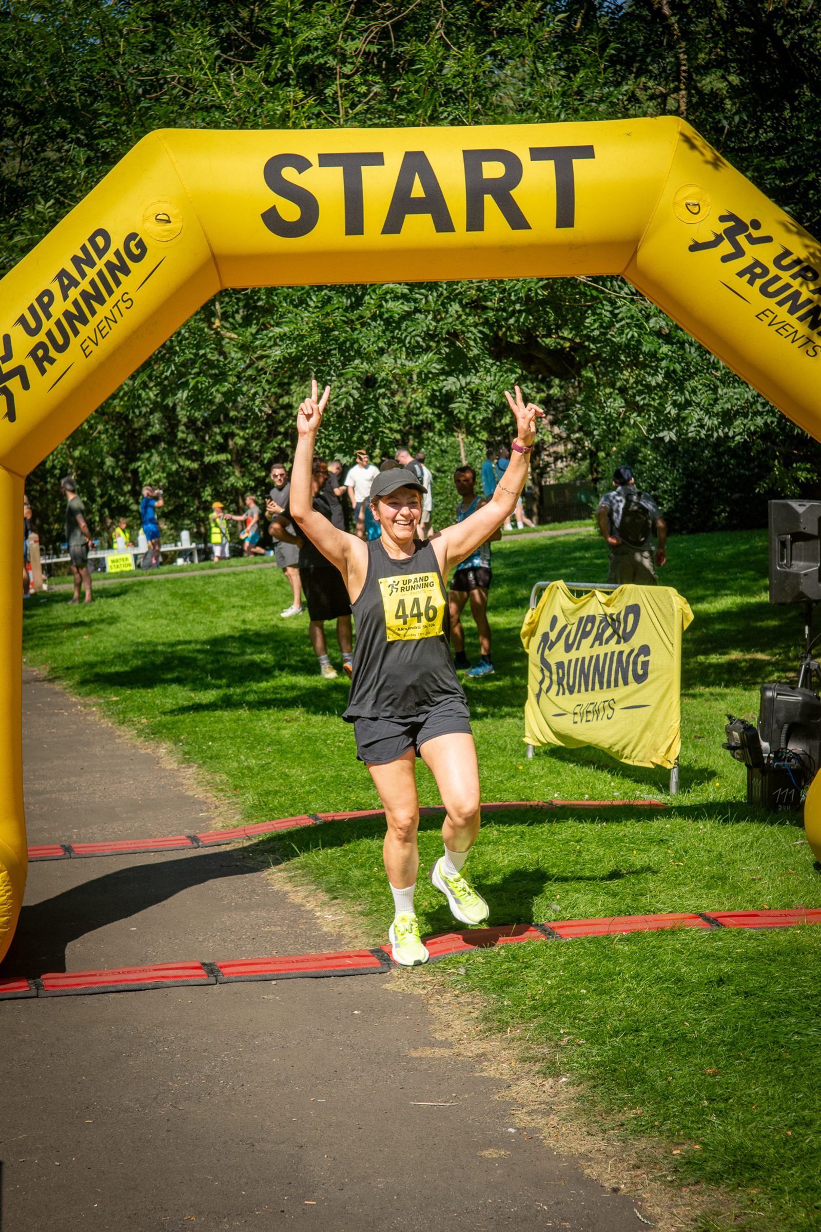 A runner wearing race bib 446 and a cap crosses the starting line under a yellow "START" arch at an outdoor event, smiling and raising both hands in a victory sign. Trees and other participants are visible in the background.