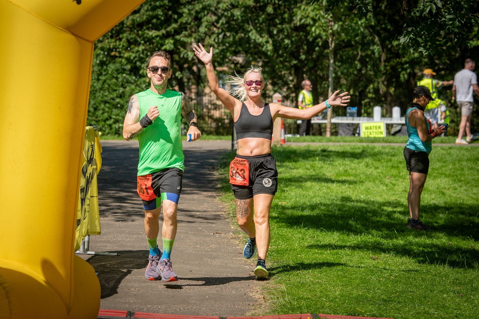 Two runners, a man in a green shirt and a woman in a black top and shorts, smile and raise their arms as they cross a race finish line outdoors in a sunny park. Other people and trees are visible in the background.
