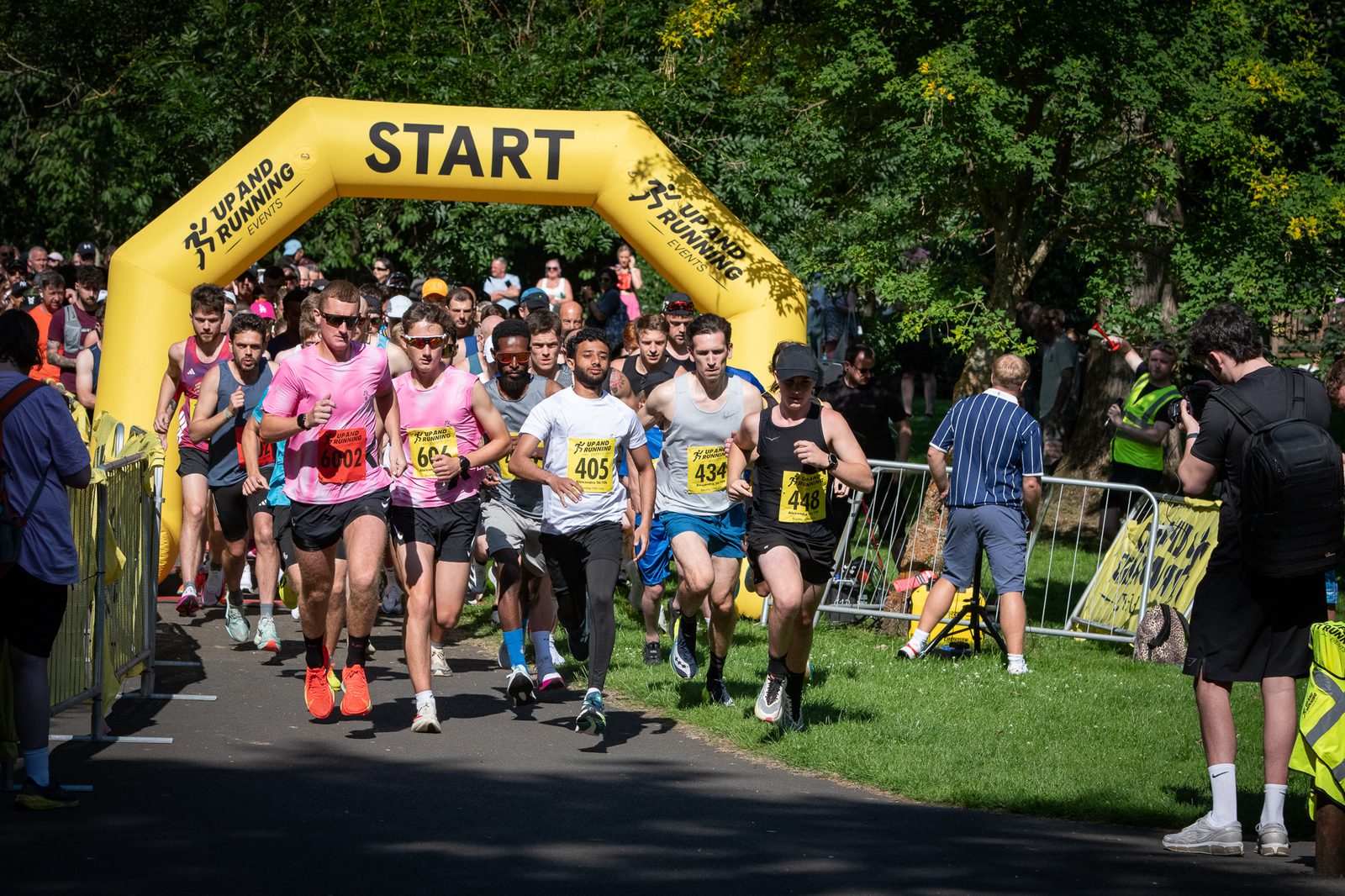 A group of runners in athletic gear begin a race under a yellow "START" arch in a park, with spectators and trees in the background.