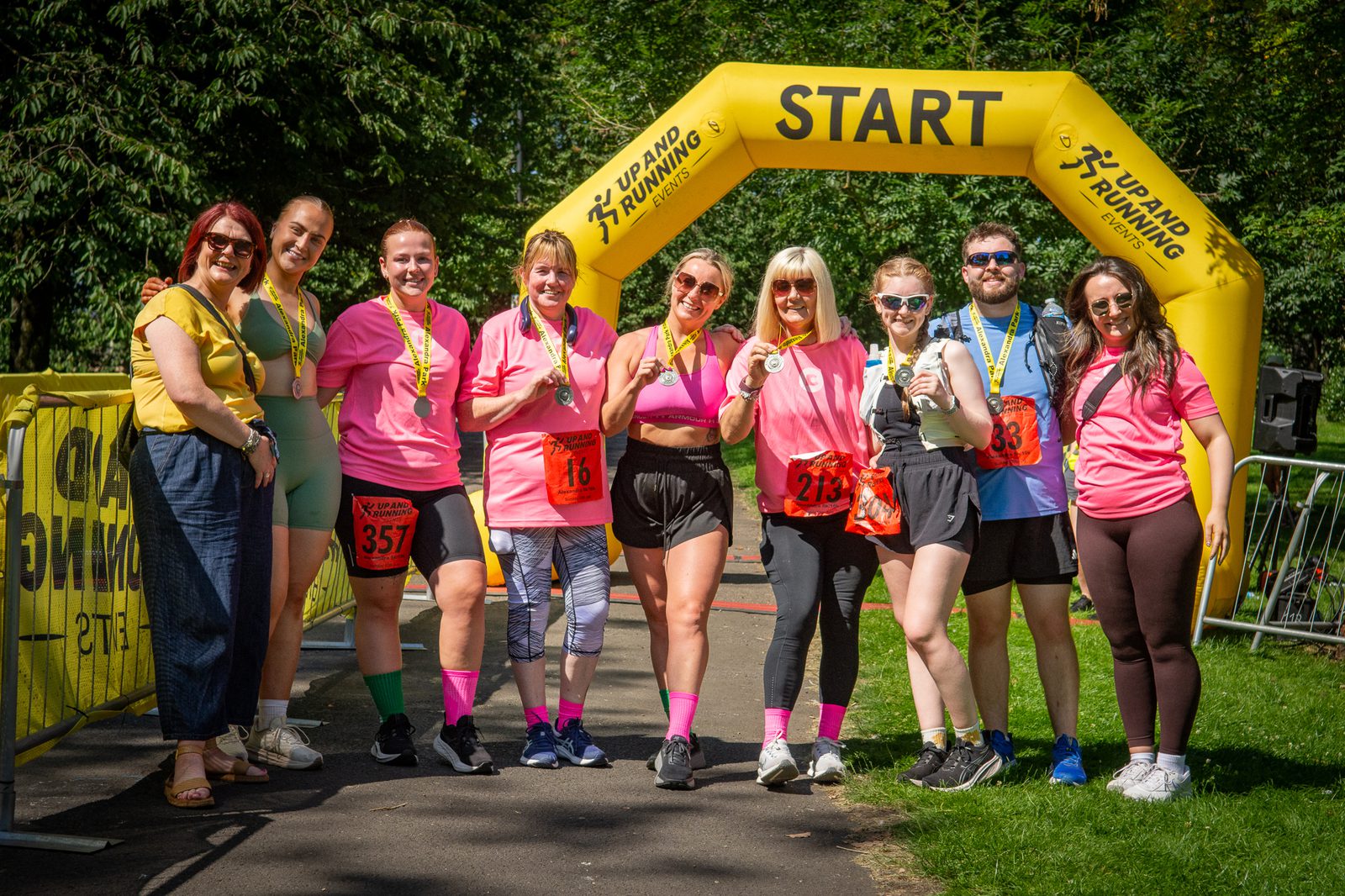 A group of nine people, some wearing matching pink shirts and medals, stand smiling under a yellow inflatable "START" arch at an outdoor running event on a sunny day.