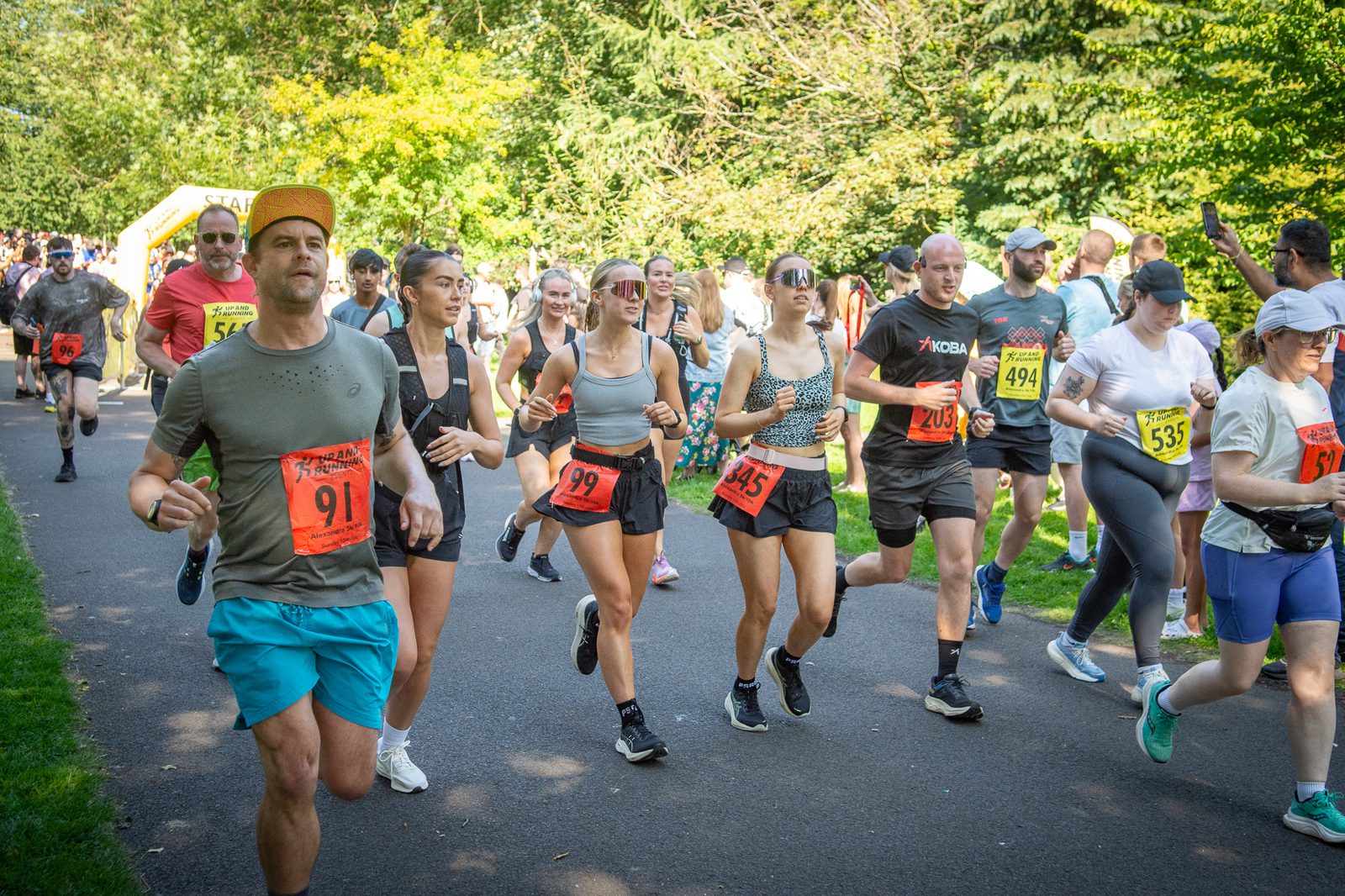 A group of people wearing numbered bibs are running and jogging on a park path during a race on a sunny day, surrounded by trees and greenery.