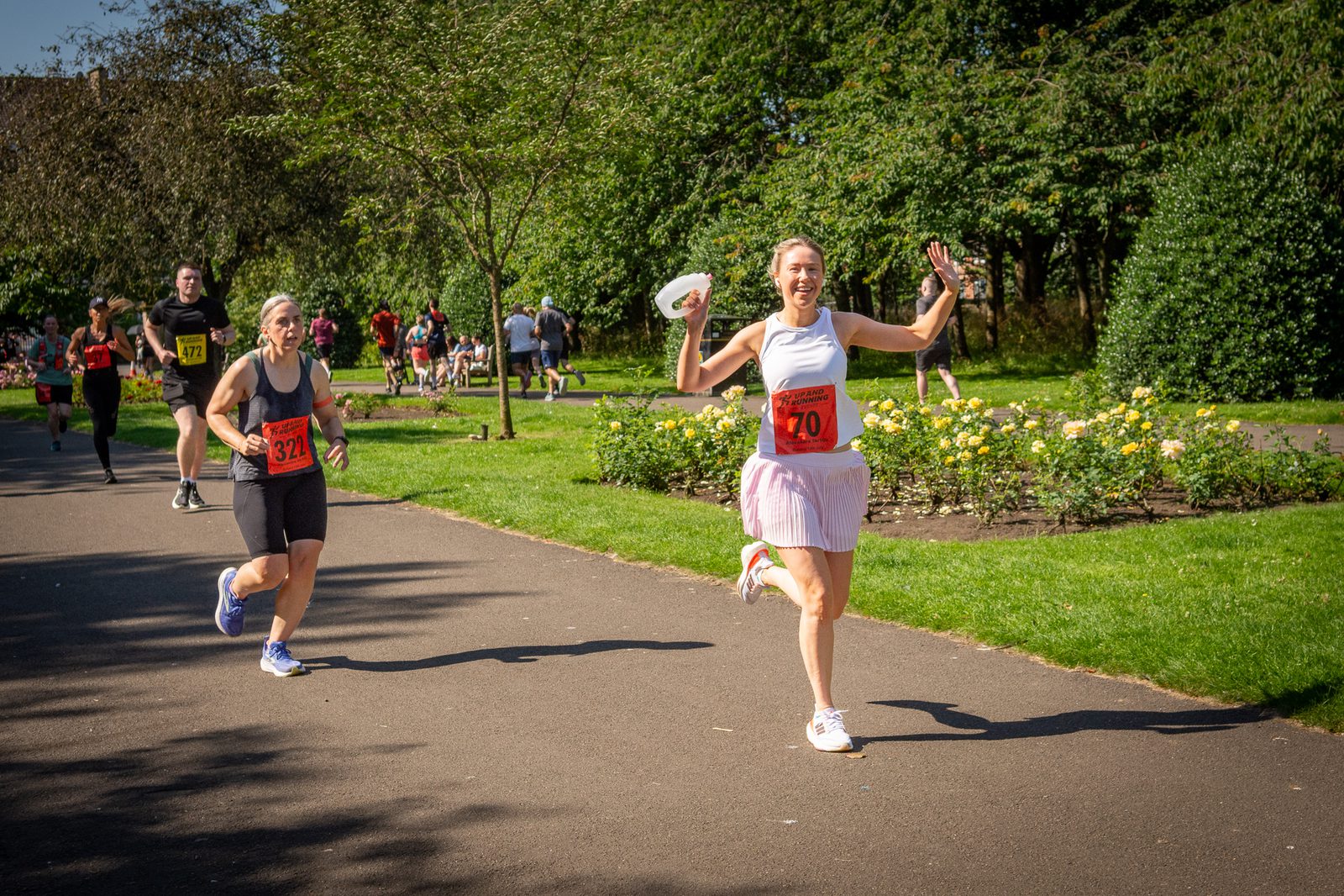 Two women are running in a race through a sunny park. The woman in front, wearing a white tank top and pink skirt, is smiling, waving, and holding a cap. Other runners and people are visible in the background.