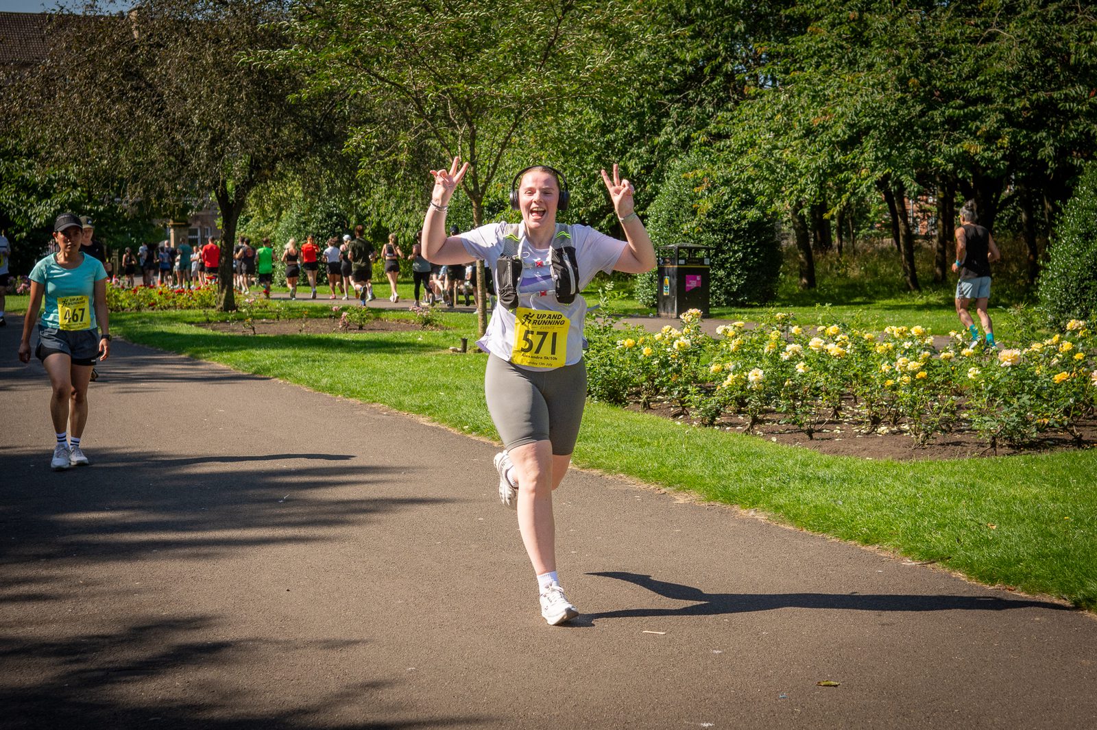 A runner wearing a race bib number 571 smiles and makes peace signs with both hands while running on a sunny park path lined with grass and flowers. Other runners and people are visible in the background.