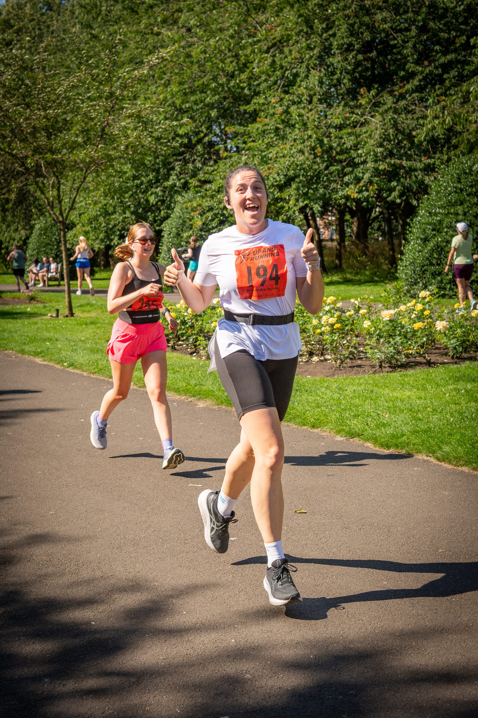 Two women are running in a race through a park. The woman in front, wearing a white shirt and black shorts, smiles and gives a thumbs-up. The second woman, in a pink skirt and orange tank top, runs behind her. Trees and flowers line the path.