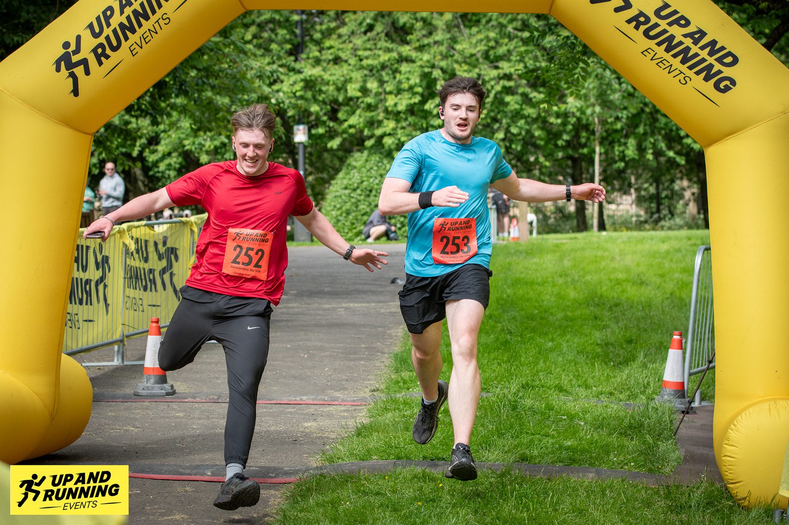 Two runners, one in a red shirt numbered 252 and the other in a blue shirt numbered 253, race each other towards a yellow inflatable archway that reads "Up And Running Events." They are sprinting on a path surrounded by greenery, reaching the finish line.