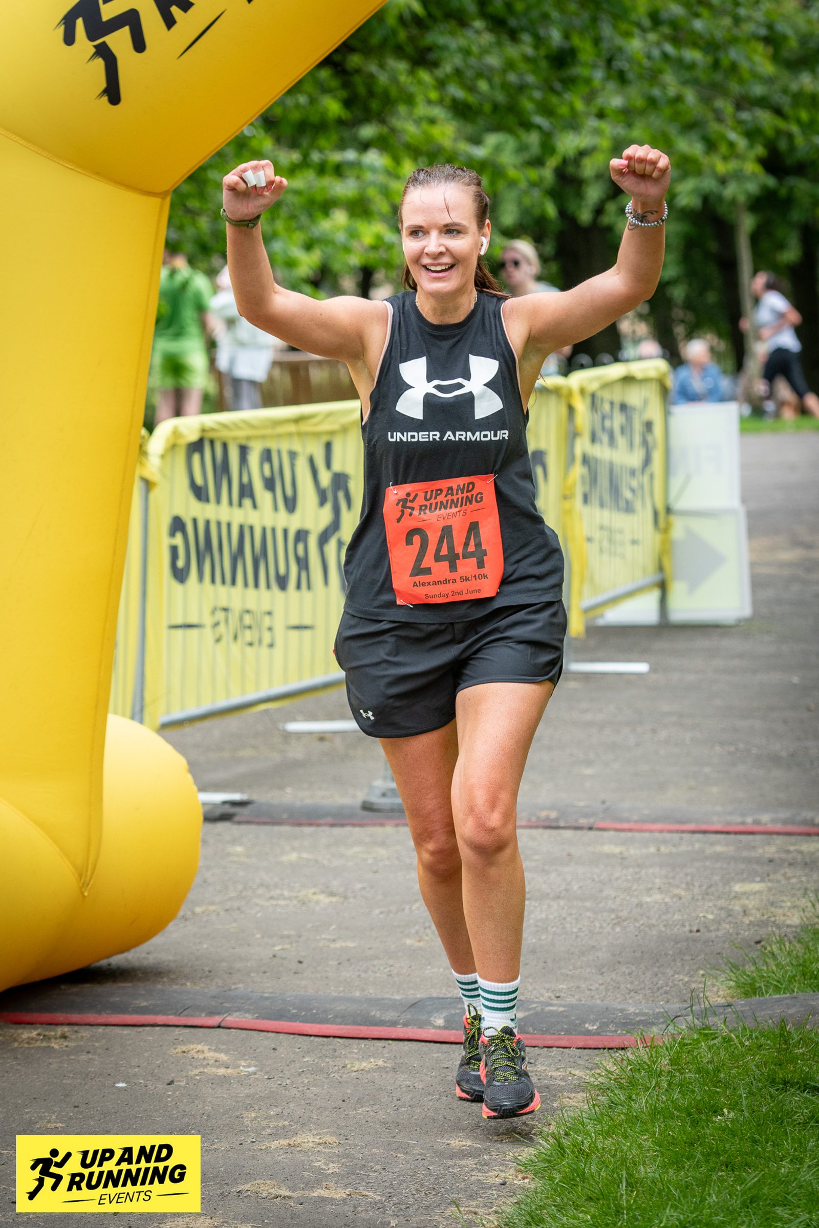 A runner wearing a black Under Armour tank top, black shorts, and bib number 244 raises both arms in celebration as she crosses the finish line of a race. There is a yellow inflatable arch and event signage behind her with trees and spectators in the background.