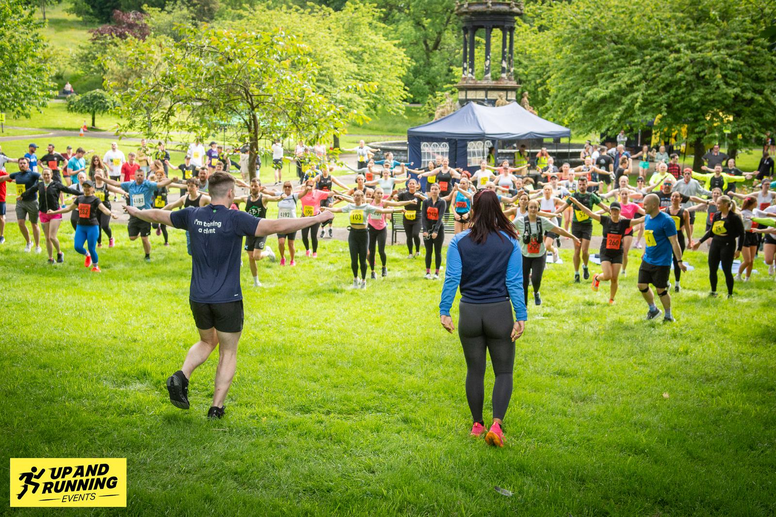 Group of people stretching in a park before a race. Two instructors are leading the warm-up, with participants spread across the grass. A canopy and trees are in the background, and a sign reads "Up and Running Events" in the foreground.