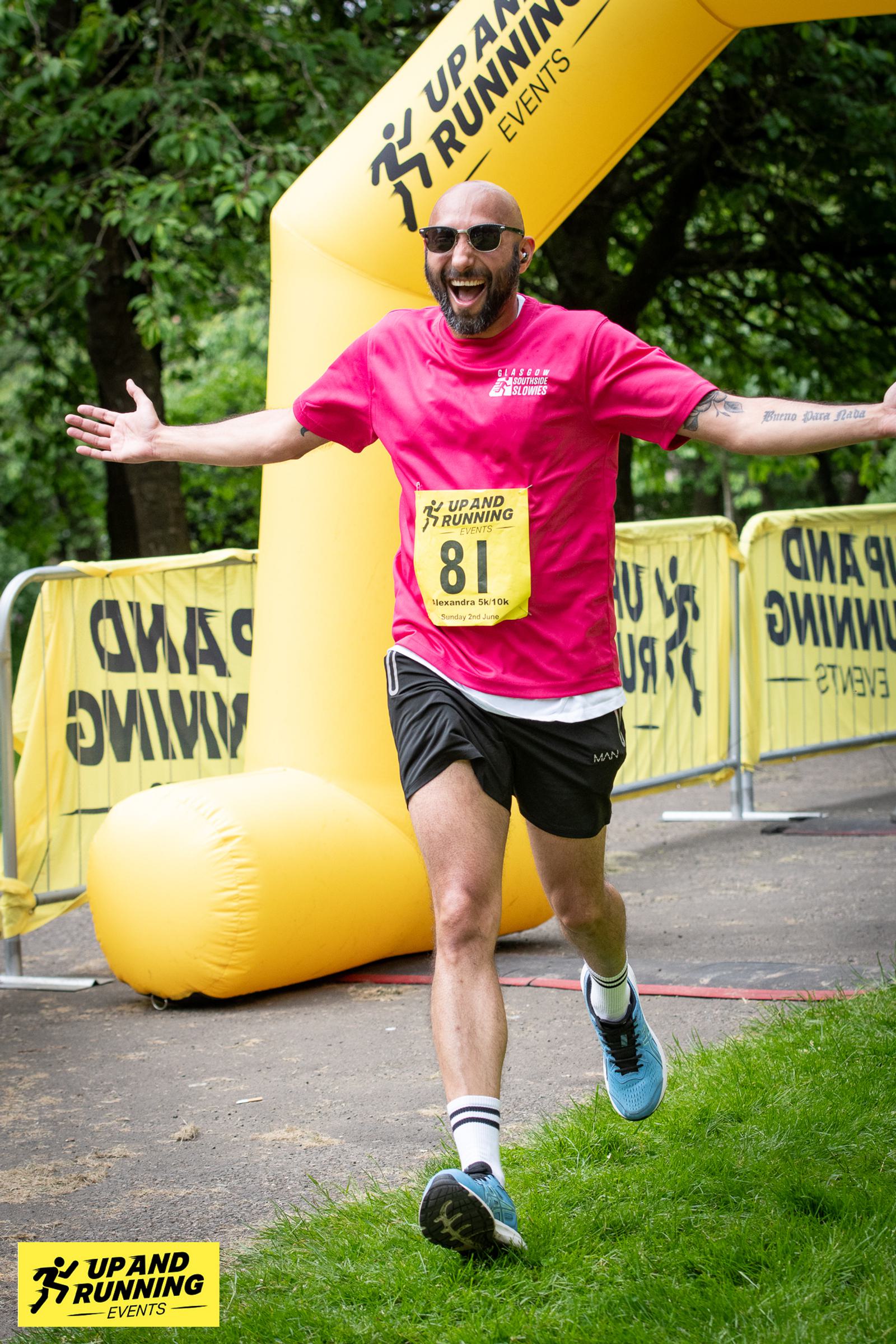 A runner wearing a bright pink shirt and black shorts joyfully crosses the finish line in a park. Behind him, a yellow inflatable arch and barriers display "Up and Running" branding. He appears to be celebrating with his arms wide open.