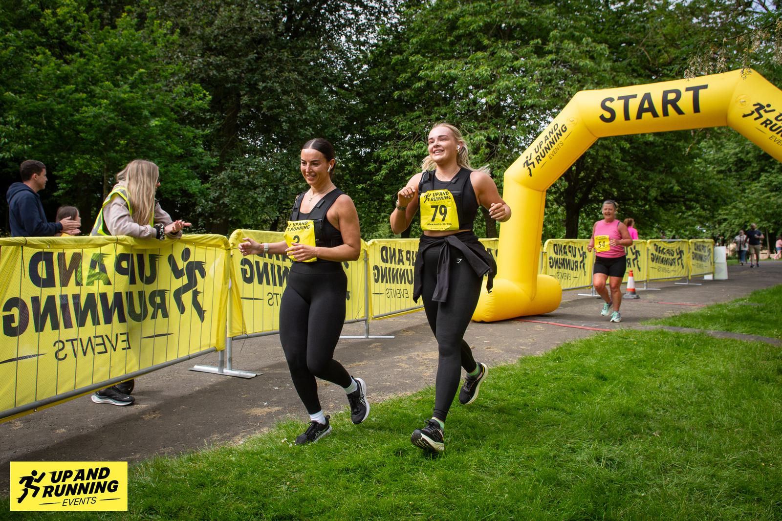 Two women in athletic wear run past a yellow "START" inflatable arch in a park setting, smiling as they participate in a race. Other runners are visible in the background. Banners display "Up and Running Events.