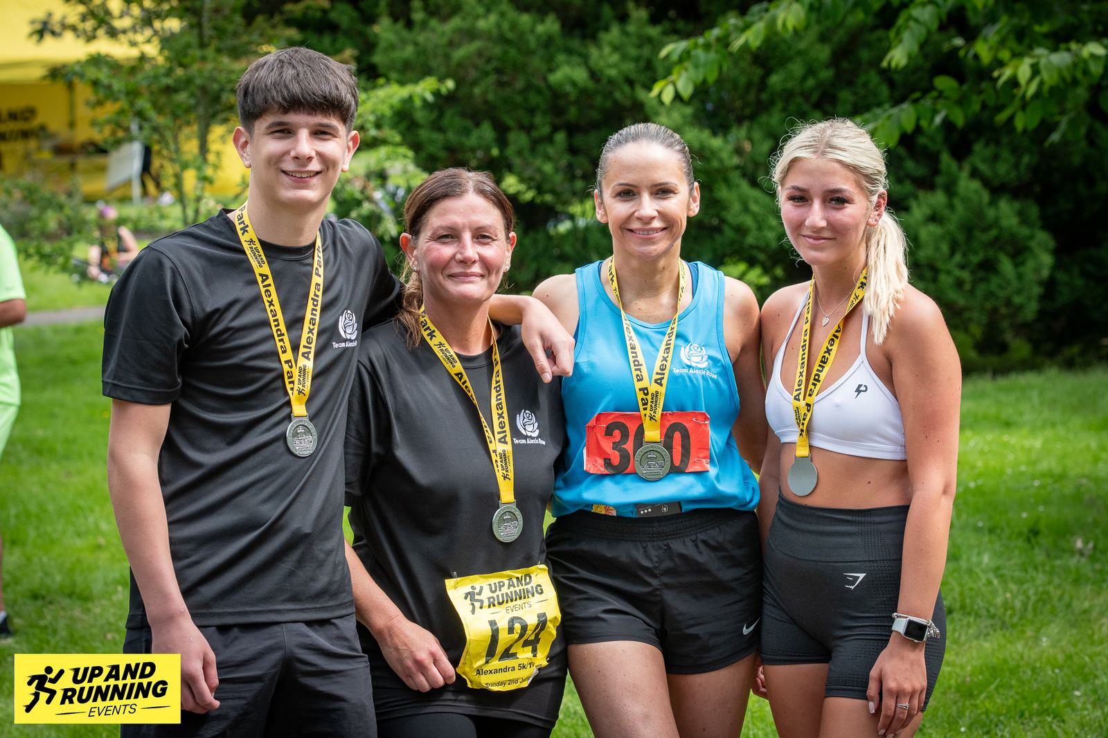 A group of four people wearing athletic gear and medals stand outdoors on grass. They are smiling at the camera, with race numbers visible on their shirts. A "Up and Running Events" sign is in the foreground. Trees are in the background.