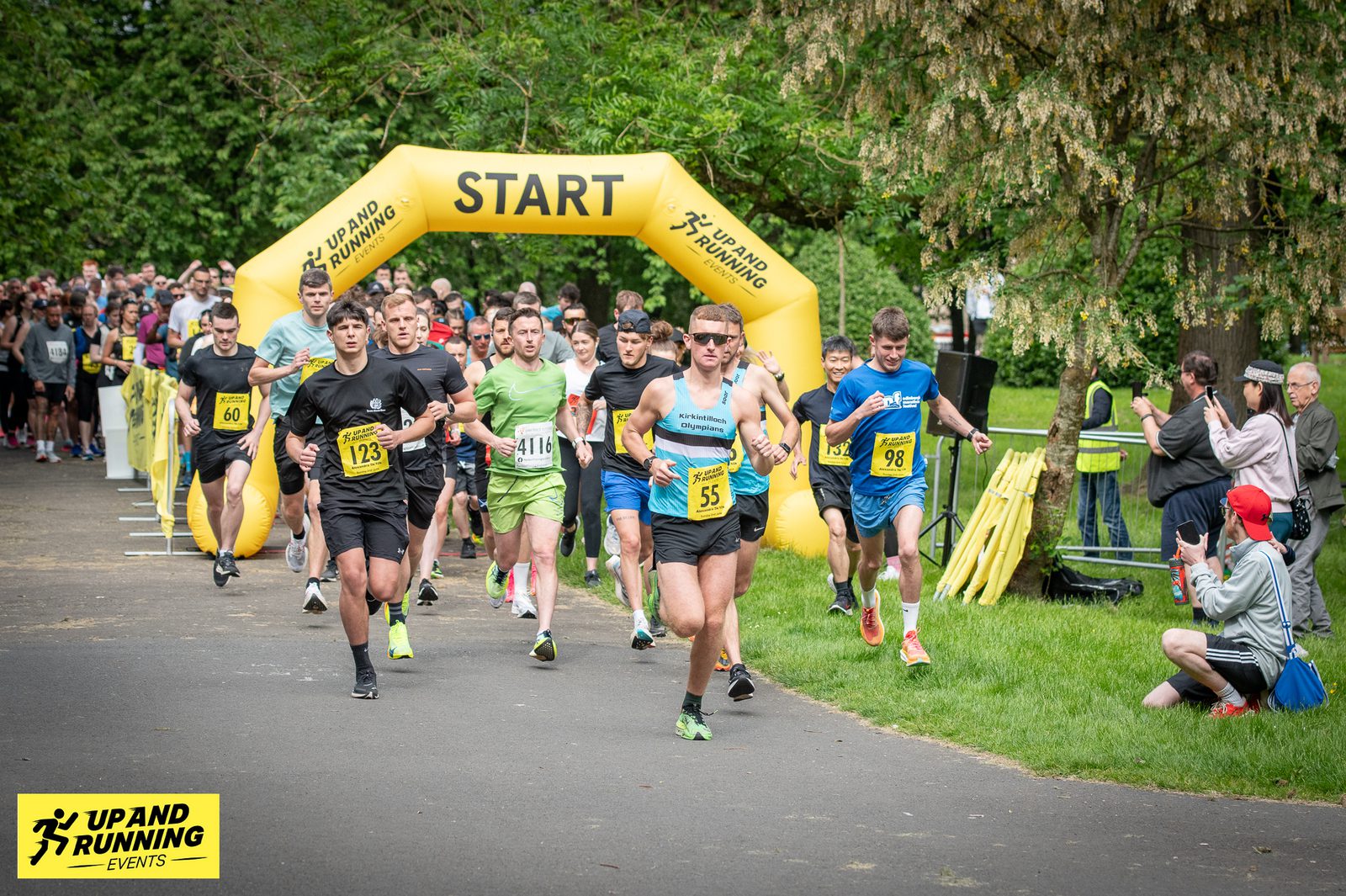 A group of runners, each wearing numbered bibs, begins a race by passing through a yellow inflatable "START" arch. The event takes place in a park with spectators watching. In the background, a banner reads "UP AND RUNNING EVENTS.