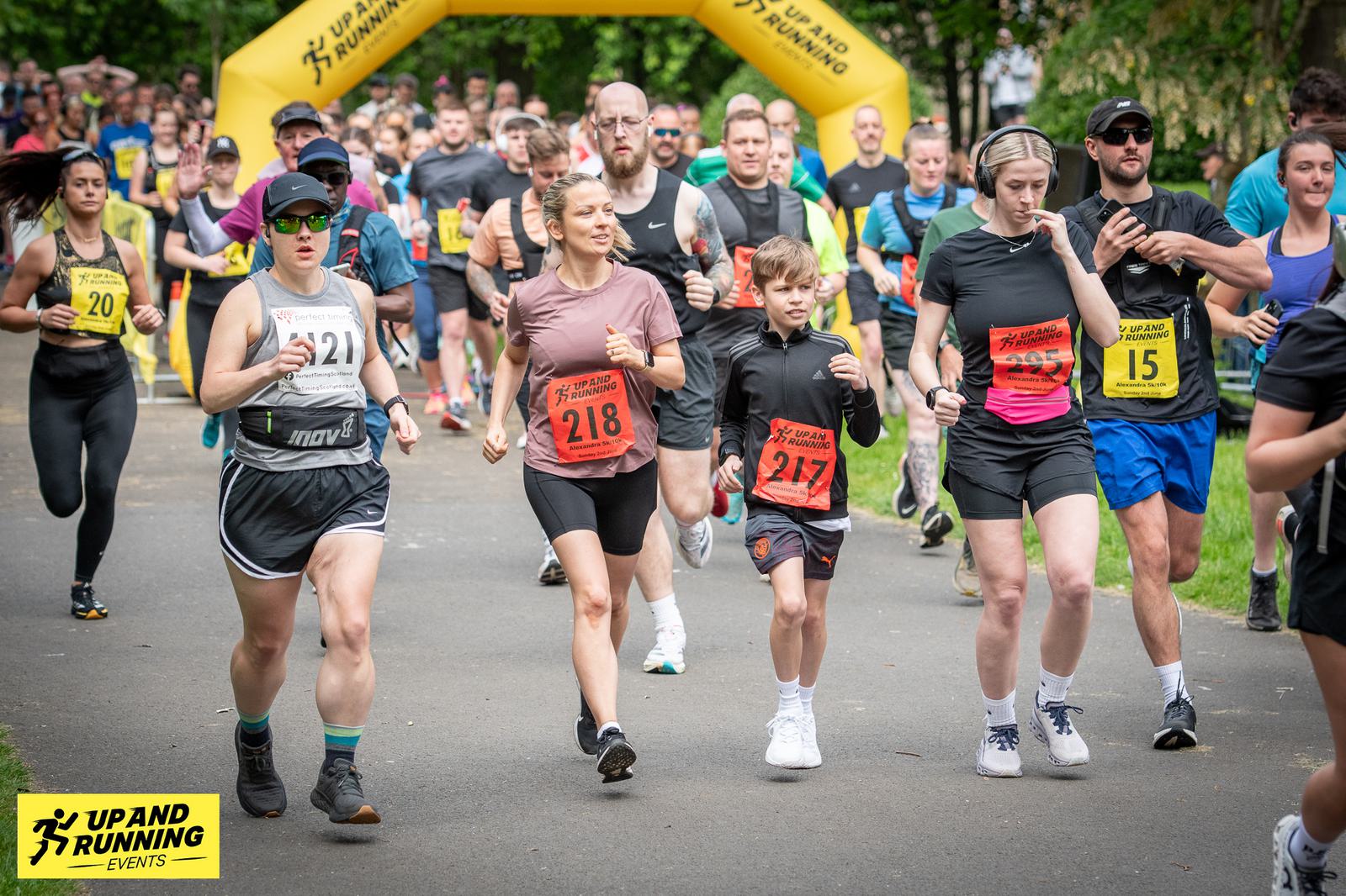 A group of runners, including women, men, and children, are participating in a race. They are wearing numbered bibs and running on a paved path. A yellow inflatable arch in the background displays "Up and Running Events.