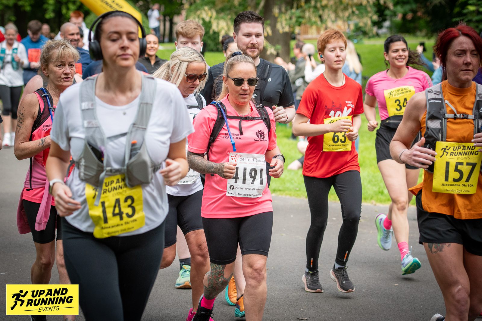 A group of runners participating in a race organized by "Up and Running Events." They are jogging along a path, some wearing bib numbers and hydration packs. Trees and grass are visible in the background, suggesting the race is taking place in a park.