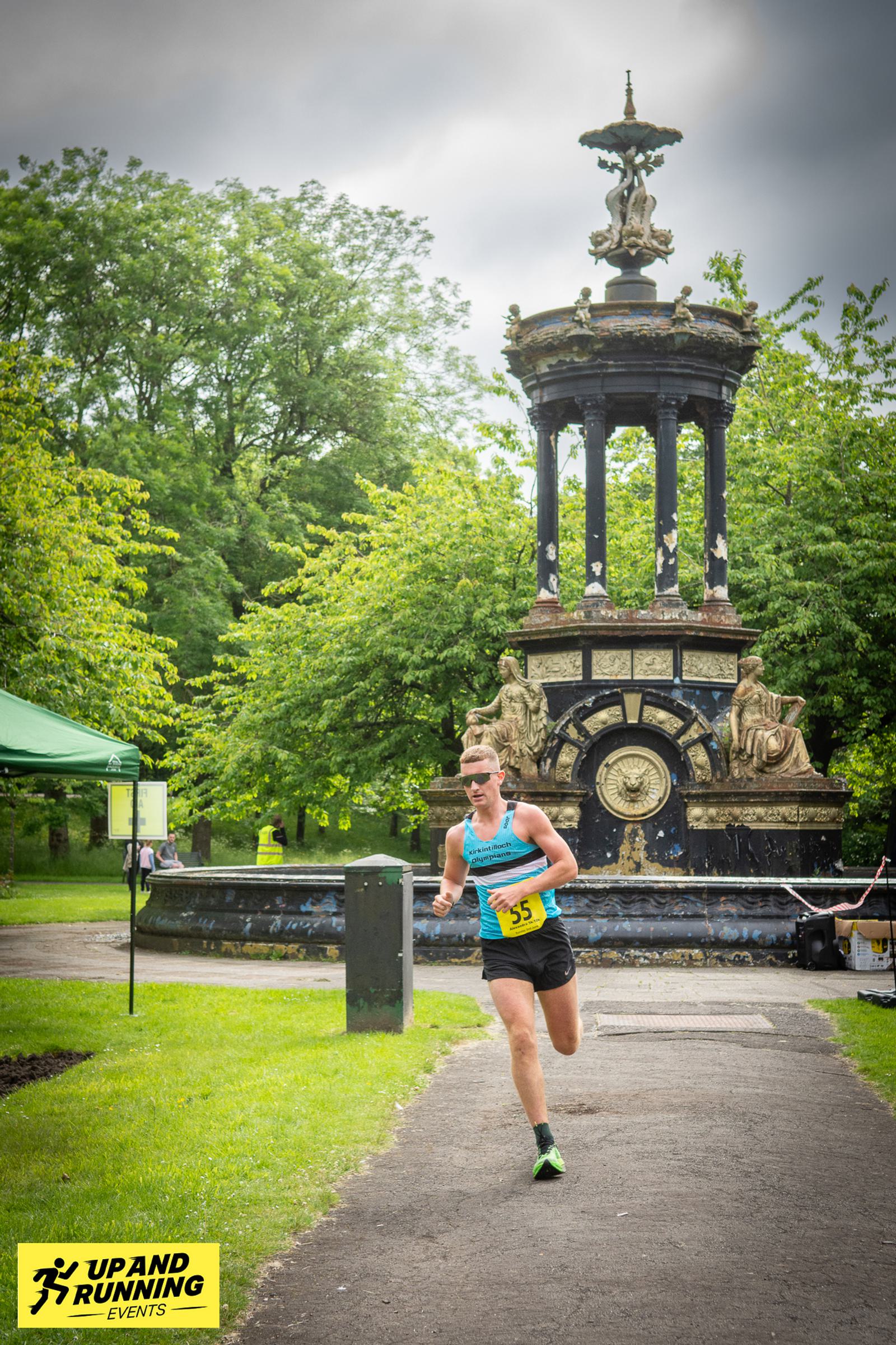 A runner in athletic gear competes in an outdoor race, passing by a decorative fountain in a park. The event is organized by Up and Running Events, as indicated by the logo at the bottom left. Trees and a cloudy sky are in the background.