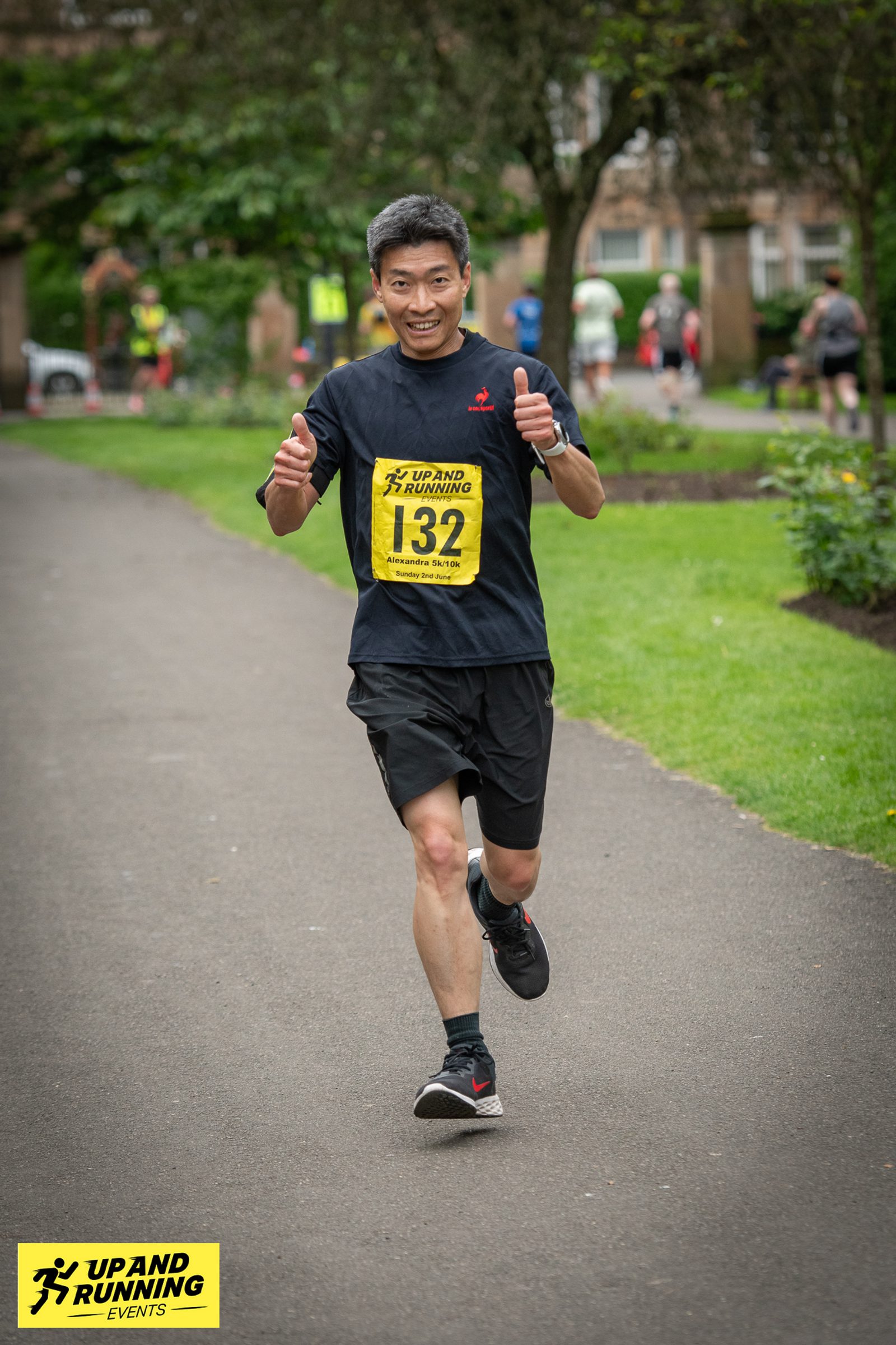 A man wearing a black shirt and shorts, with a yellow race bib numbered 132, is running on a park path during an event hosted by Up and Running Events. He is giving a thumbs-up with both hands and smiling. Trees and greenery are visible in the background.