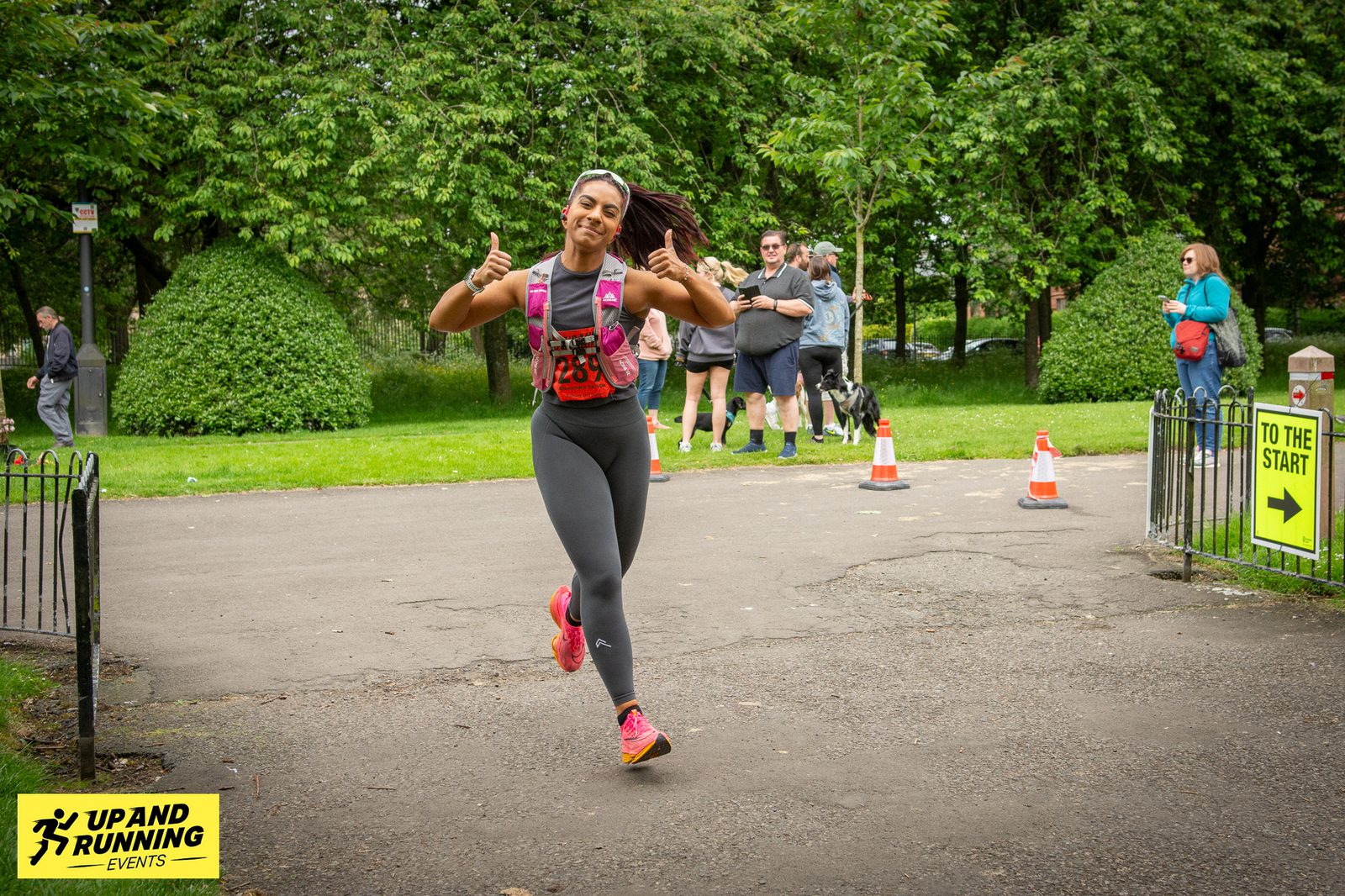 A person in athletic gear is running in a park, giving a thumbs-up with both hands. Other people and a dog are seen in the background. There are signs indicating "TO THE START." The event logo "UP & RUNNING EVENTS" is visible at the bottom left corner.