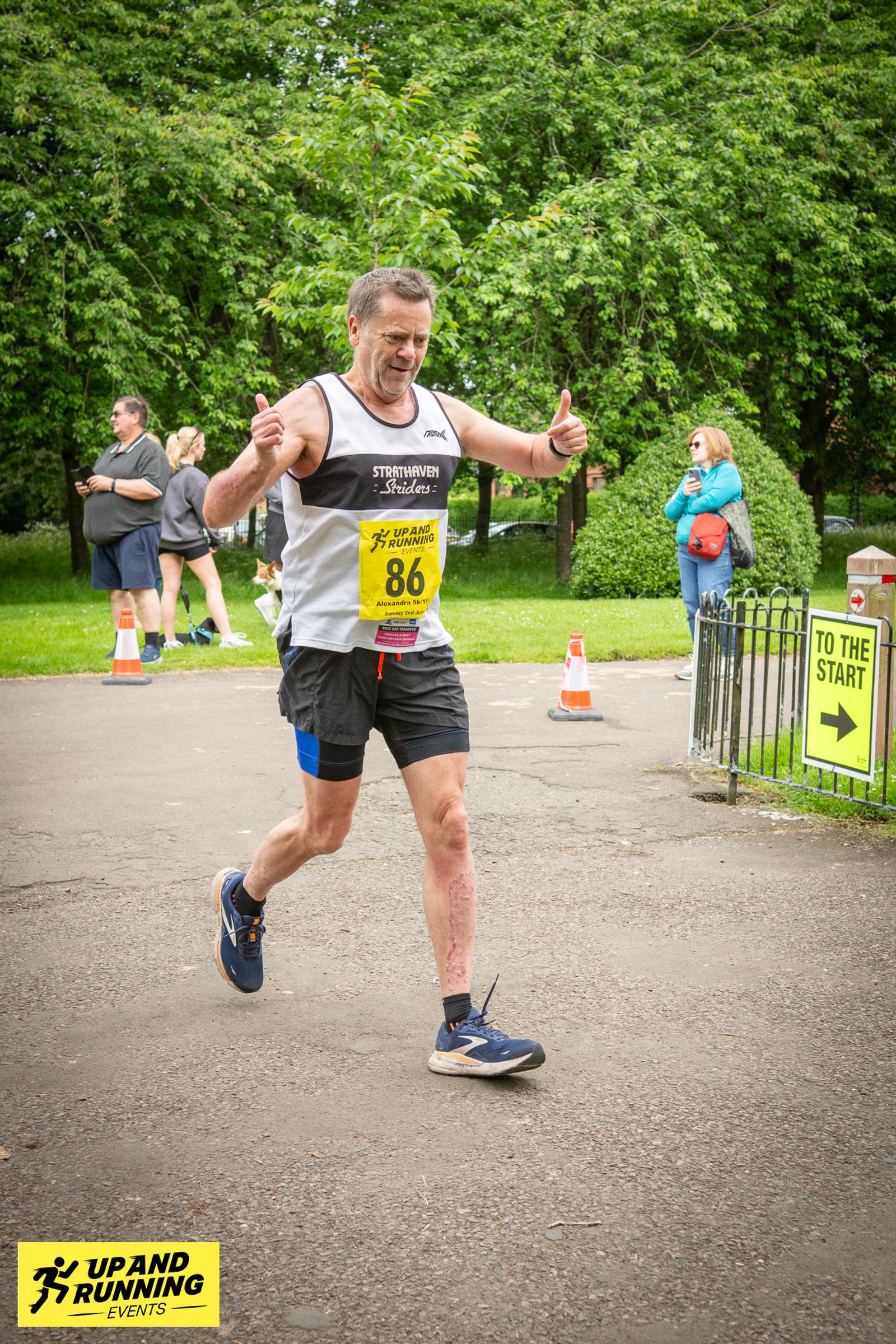 A runner wearing a white and black tank top and black shorts gives a thumbs-up as he nears the finish line in a race. He's wearing a yellow bib numbered 86. Spectators and trees are in the background, and signs indicate the path to the start.