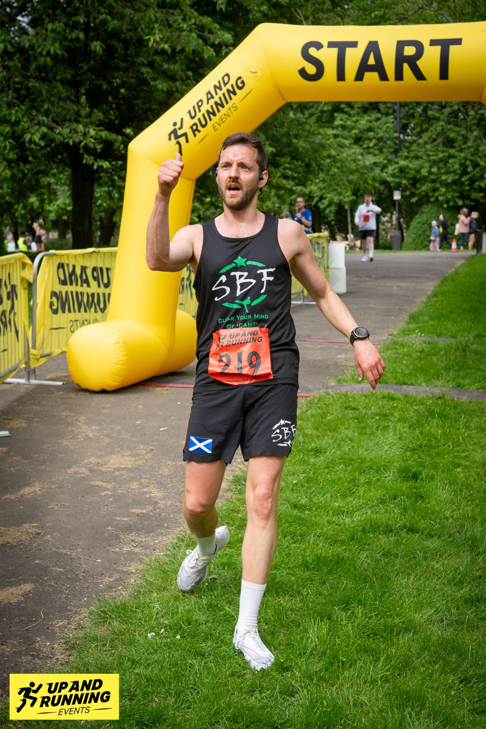 A runner wearing a black vest finishes a race, giving a thumbs-up gesture. He passes under a yellow "START" arch with "UP AND RUNNING EVENTS" branding. Trees and spectators are visible in the background.