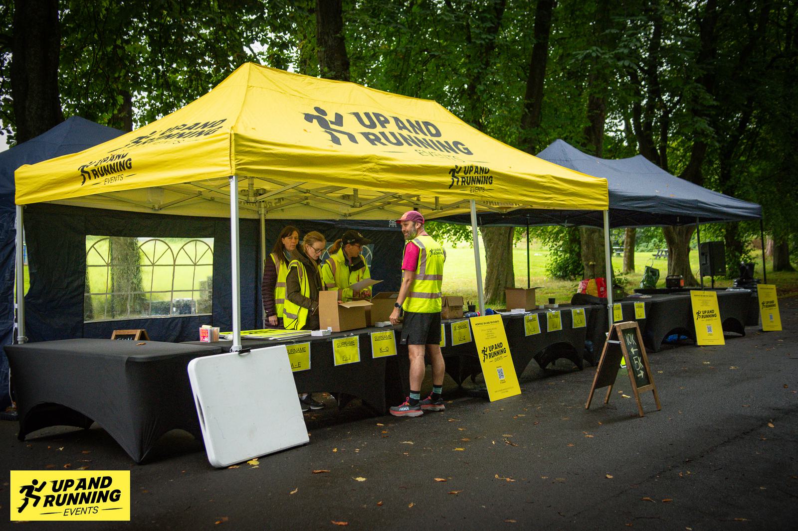 Volunteers in yellow vests gather under a yellow "Up and Running" tent, surrounded by tables and equipment, in a green park setting.