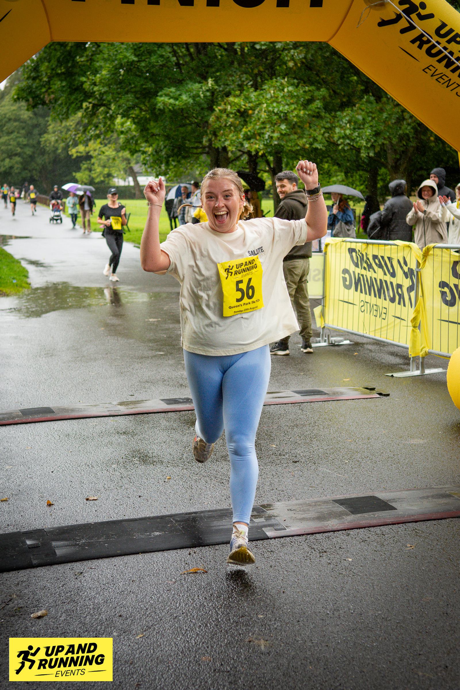 A person in a white shirt and light blue leggings joyfully crosses a race finish line under a yellow arch. They are holding their arms up in celebration. The path is wet, and spectators stand behind barriers, some with umbrellas.