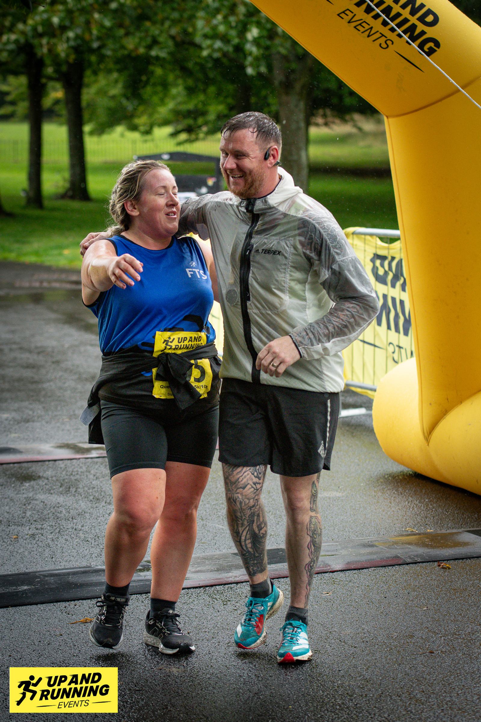 Two people wearing athletic gear cross a finish line together, smiling and embracing. A yellow inflatable arch in the background marks the event's end. The ground is wet, suggesting rain. A "Up and Running" banner is visible on the left.