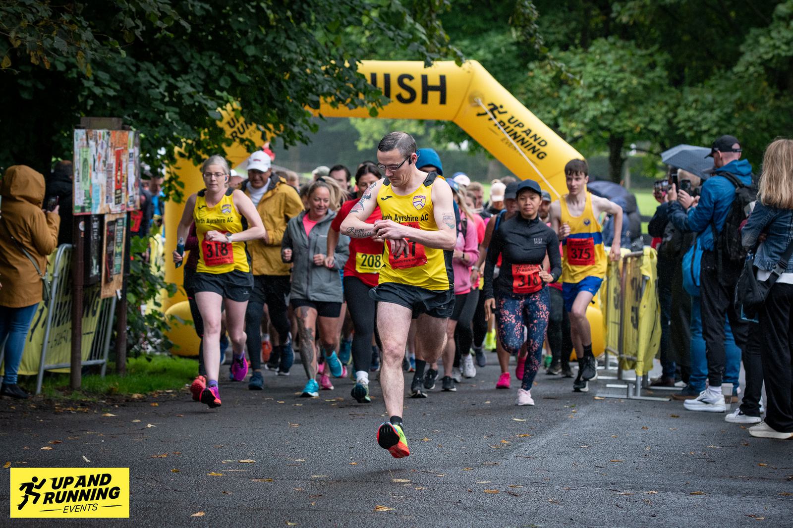A group of runners begins a race under a yellow inflatable arch labeled "FINISH" in a park setting. Participants wear race bibs and athletic gear. Spectators with umbrellas can be seen on the sidelines. A logo reads "Up and Running Events.