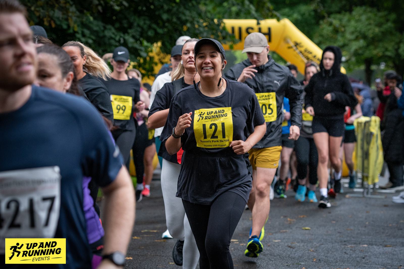 Runners participating in an outdoor event under cloudy skies. A young woman in a black shirt, wearing a race bib numbered 121, smiles as she runs. Other participants and a yellow finish arch are visible in the background.