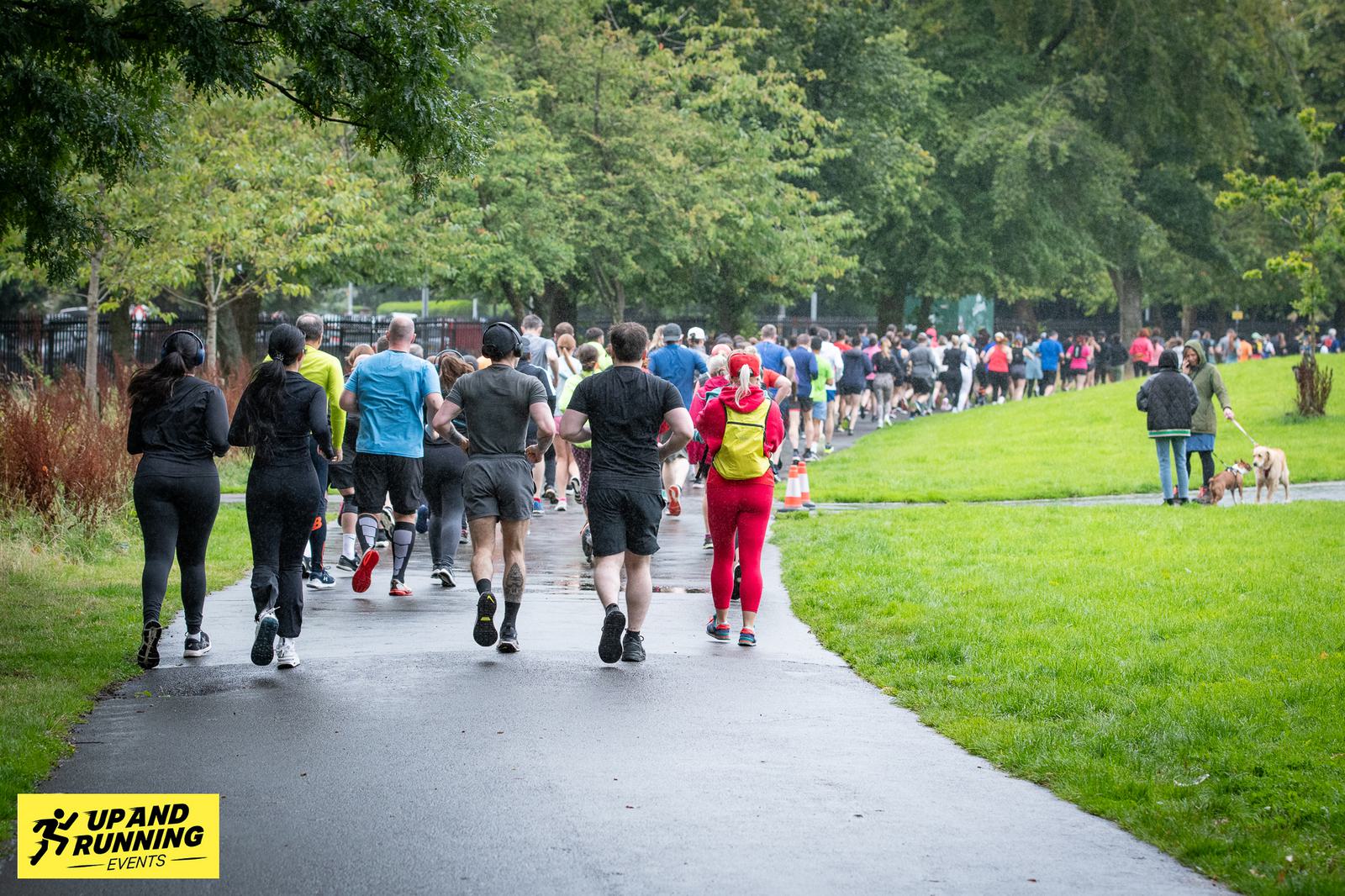 A large group of people are running along a park path during a race event. They are dressed in various athletic gear, and the path is surrounded by green grass and trees. A sign for "Up and Running Events" is visible in the foreground.