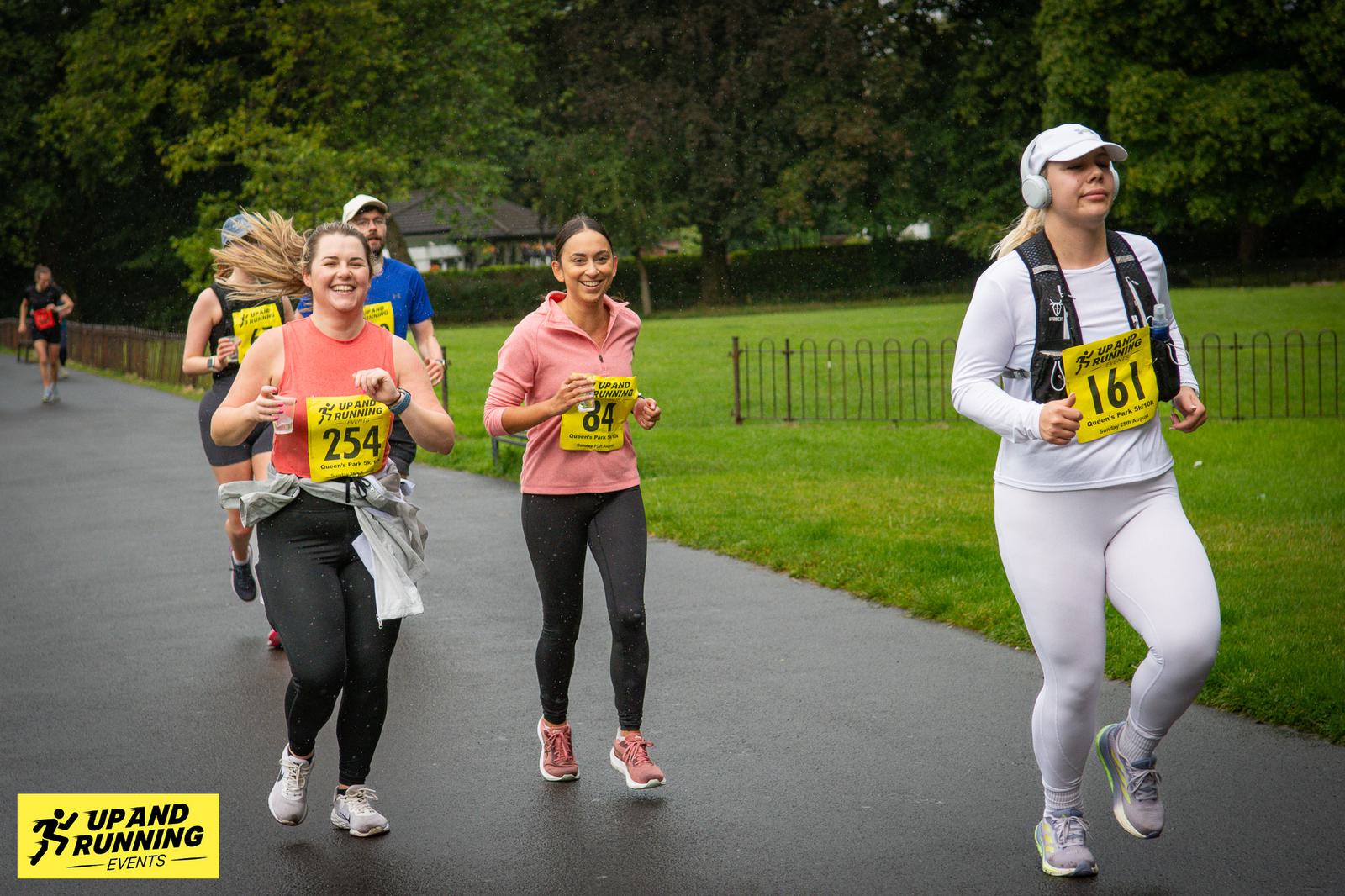 A group of runners participating in a race at a park. They are wearing athletic clothing and numbered bibs. The weather is overcast, and the ground appears wet. There's a yellow event sign in the foreground.