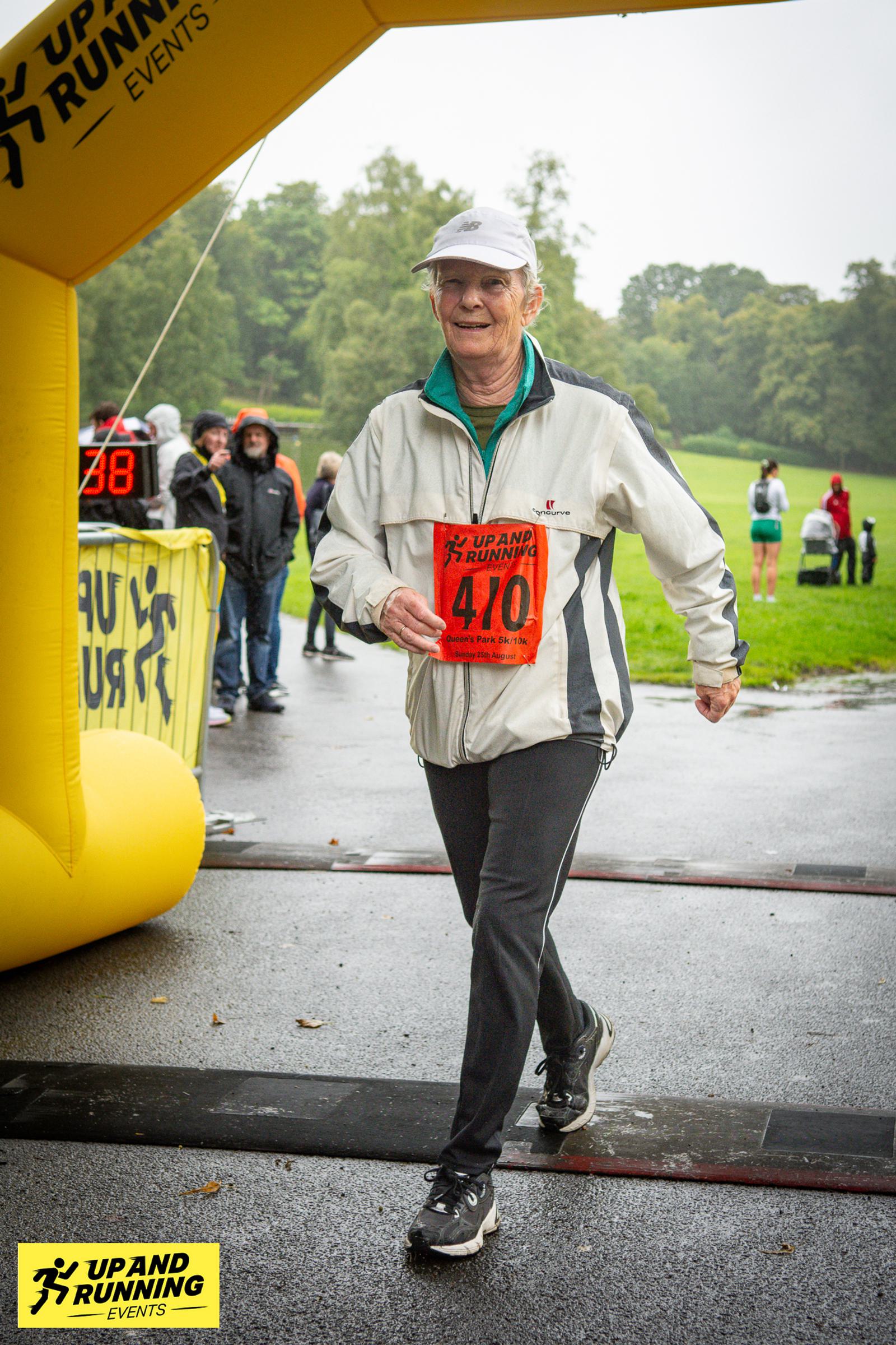 An elderly participant crossing the finish line at a running event, wearing a white jacket and cap, holding a race number 470. People are gathered in the background under overcast skies. A yellow "Up and Running" event arch is visible.