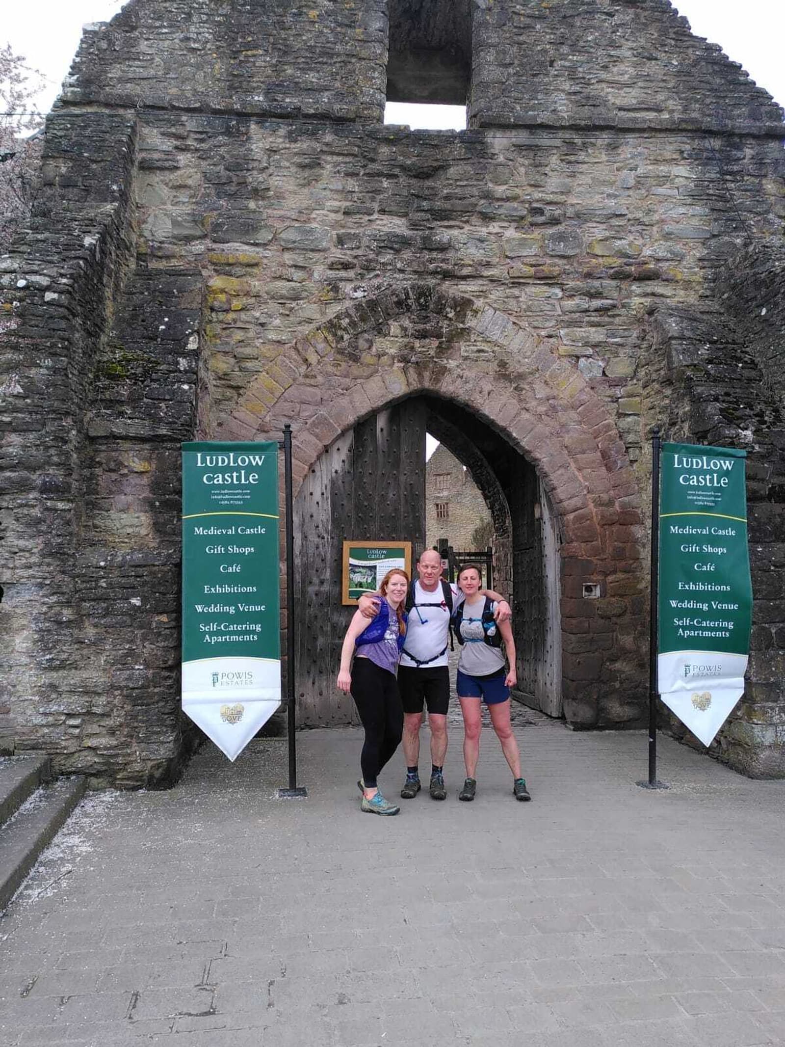 Three people pose smiling in front of the stone entrance gate to Ludlow Castle. Two green signs list visitor information about the castle on either side of the gateway. The castle archway and weathered walls are prominent.