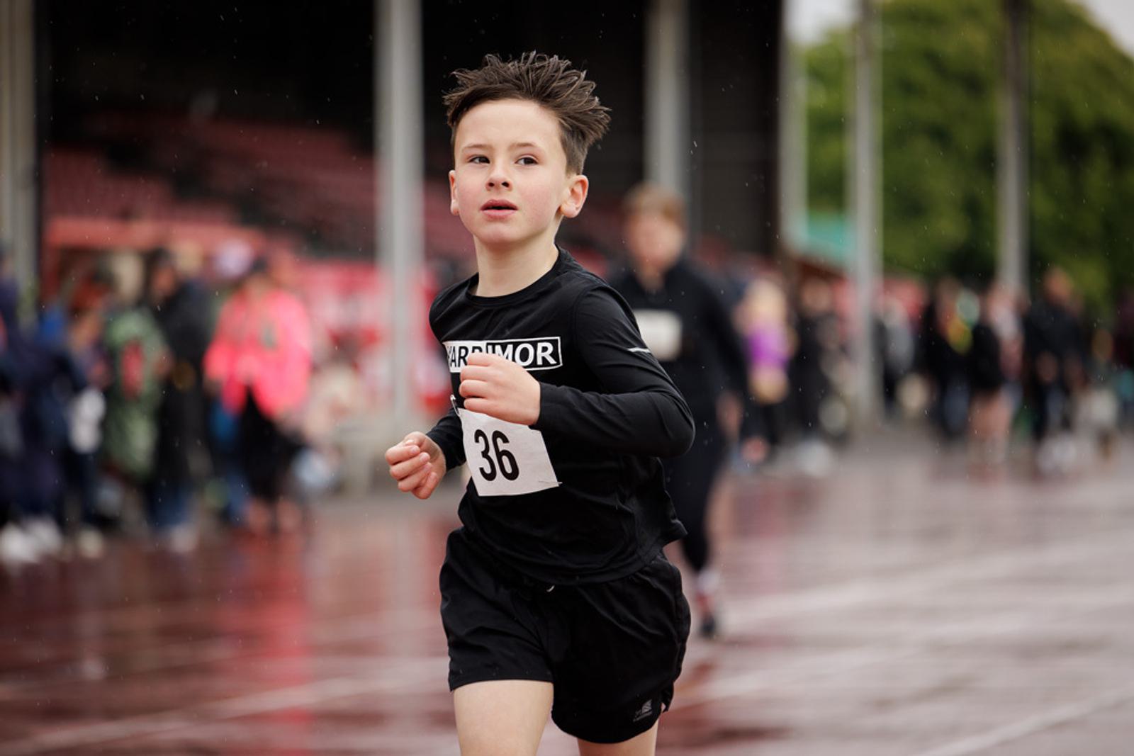 A young boy with short hair is running on a wet track, wearing a black long-sleeve shirt with a number "36" bib. The background shows blurred spectators and trees, indicating a sporting event in progress.